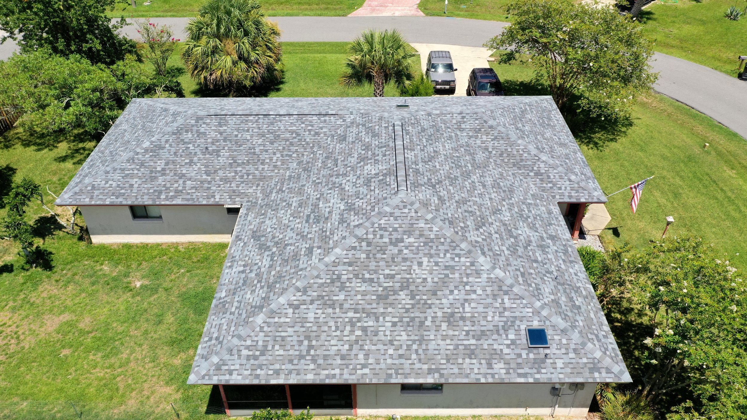 An aerial view of a house with a gray shingle roof, green lawn, trees, a driveway with parked cars, a flag on a pole, and a curved street in a neighborhood.
