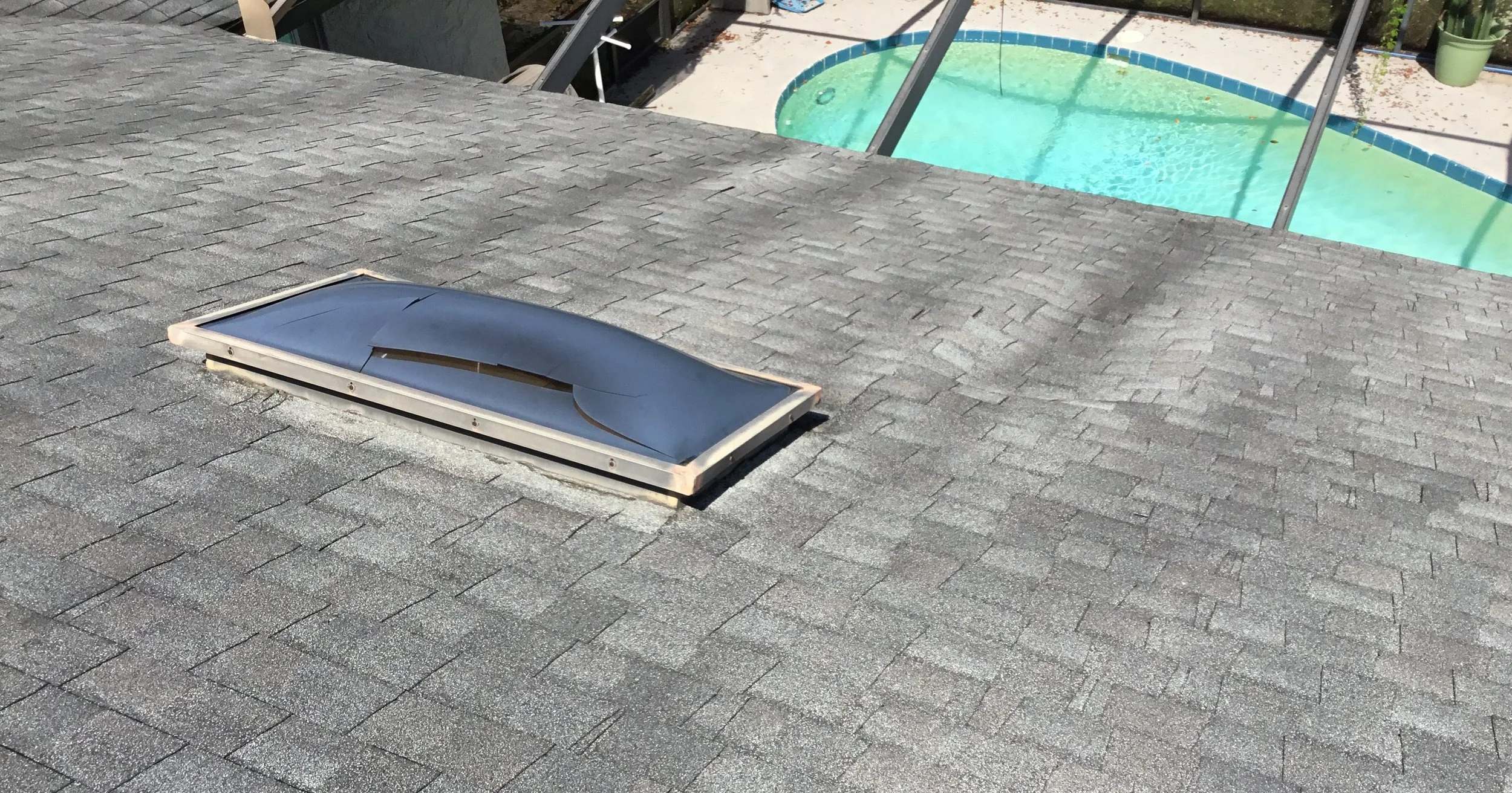View of a gray shingled roof with a damaged skylight and a backyard with a swimming pool and patio area in the background.
