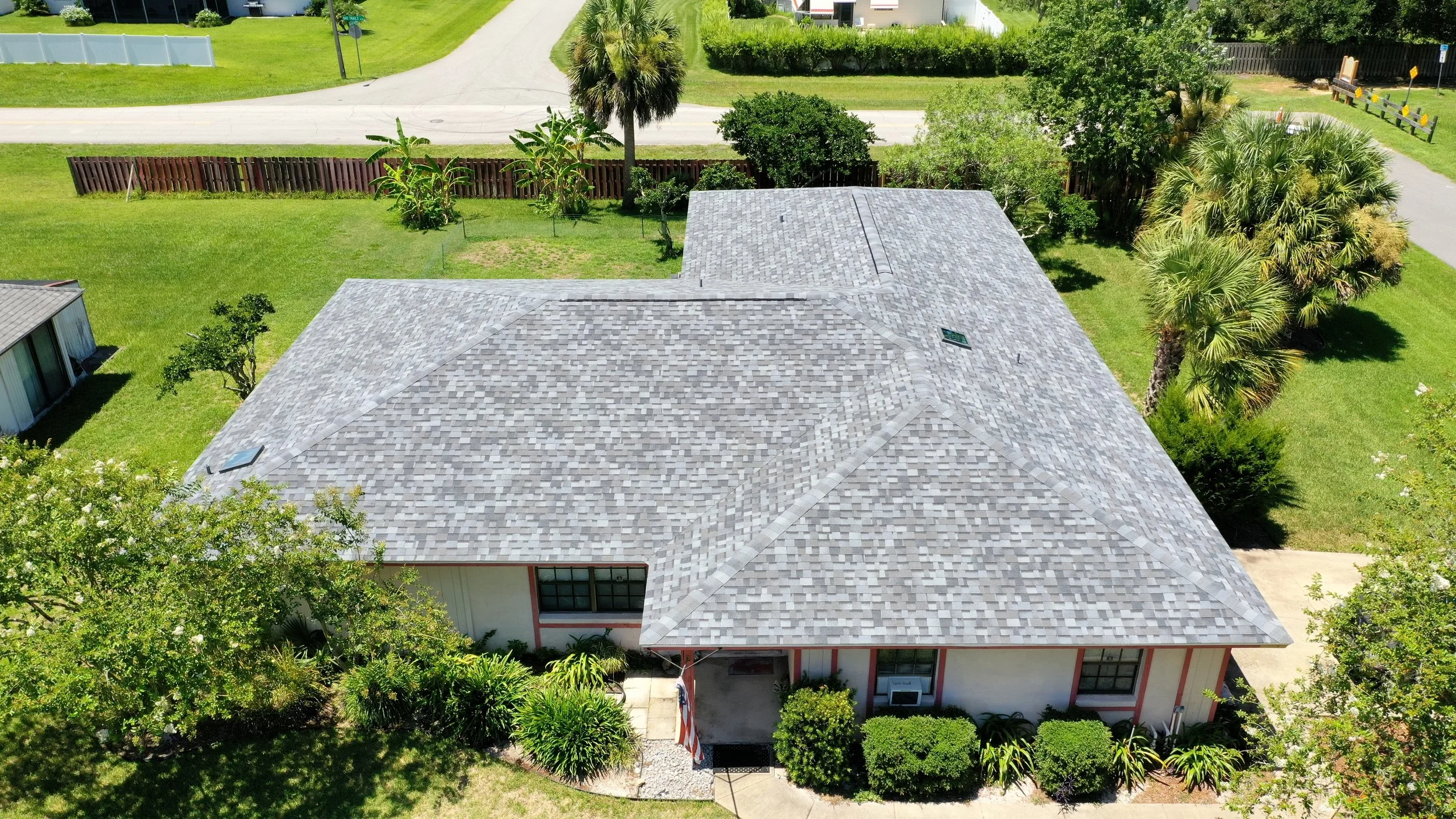 Aerial view of a single-story house with a gray shingle roof, surrounded by green lawn, trees, and shrubs, with a driveway on the right and street in the background.
