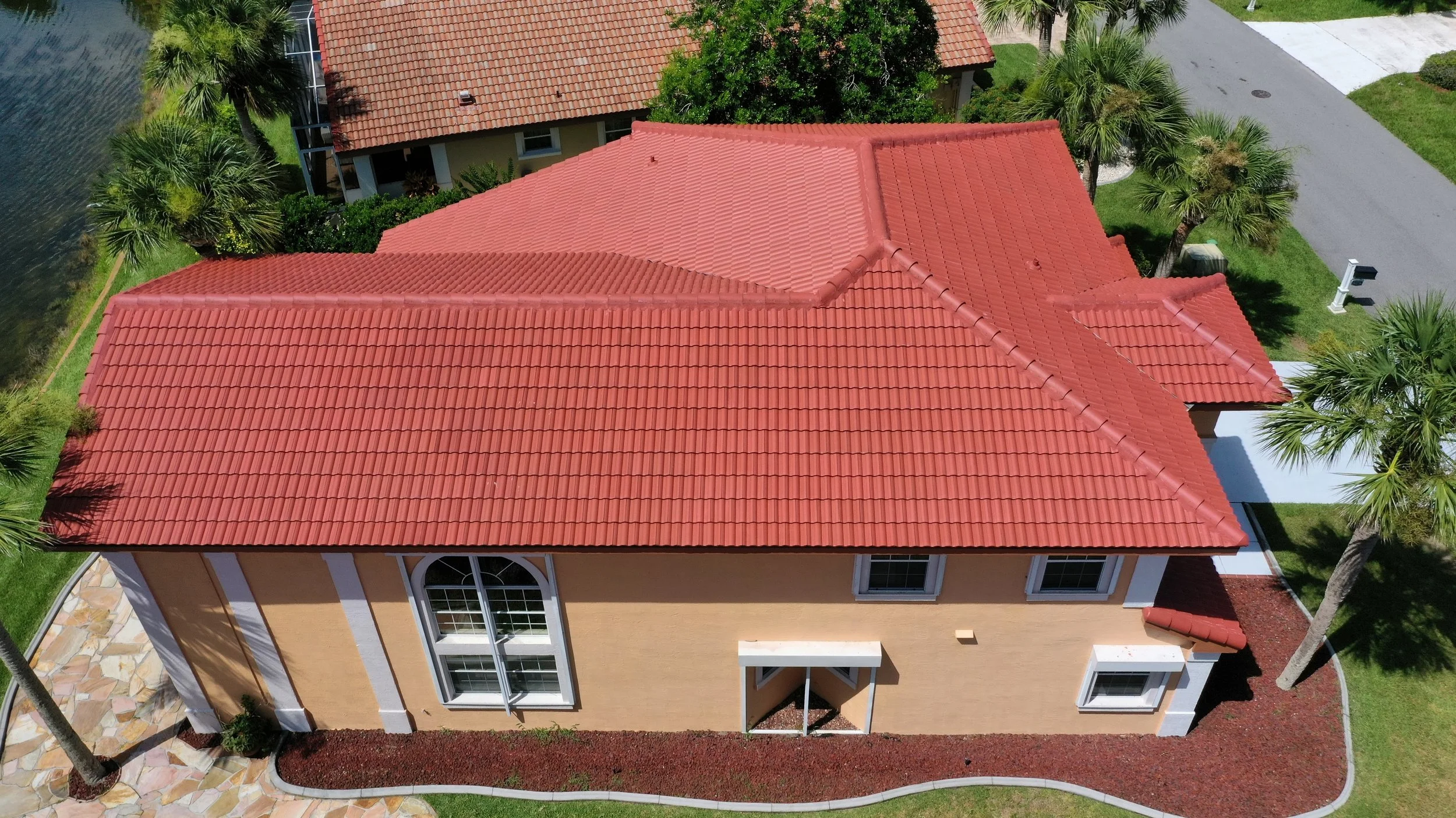 Aerial view of a house with a red tiled roof, beige exterior walls, white window frames, and a stone pathway leading to the entrance, surrounded by green grass, palm trees, and neighboring houses.