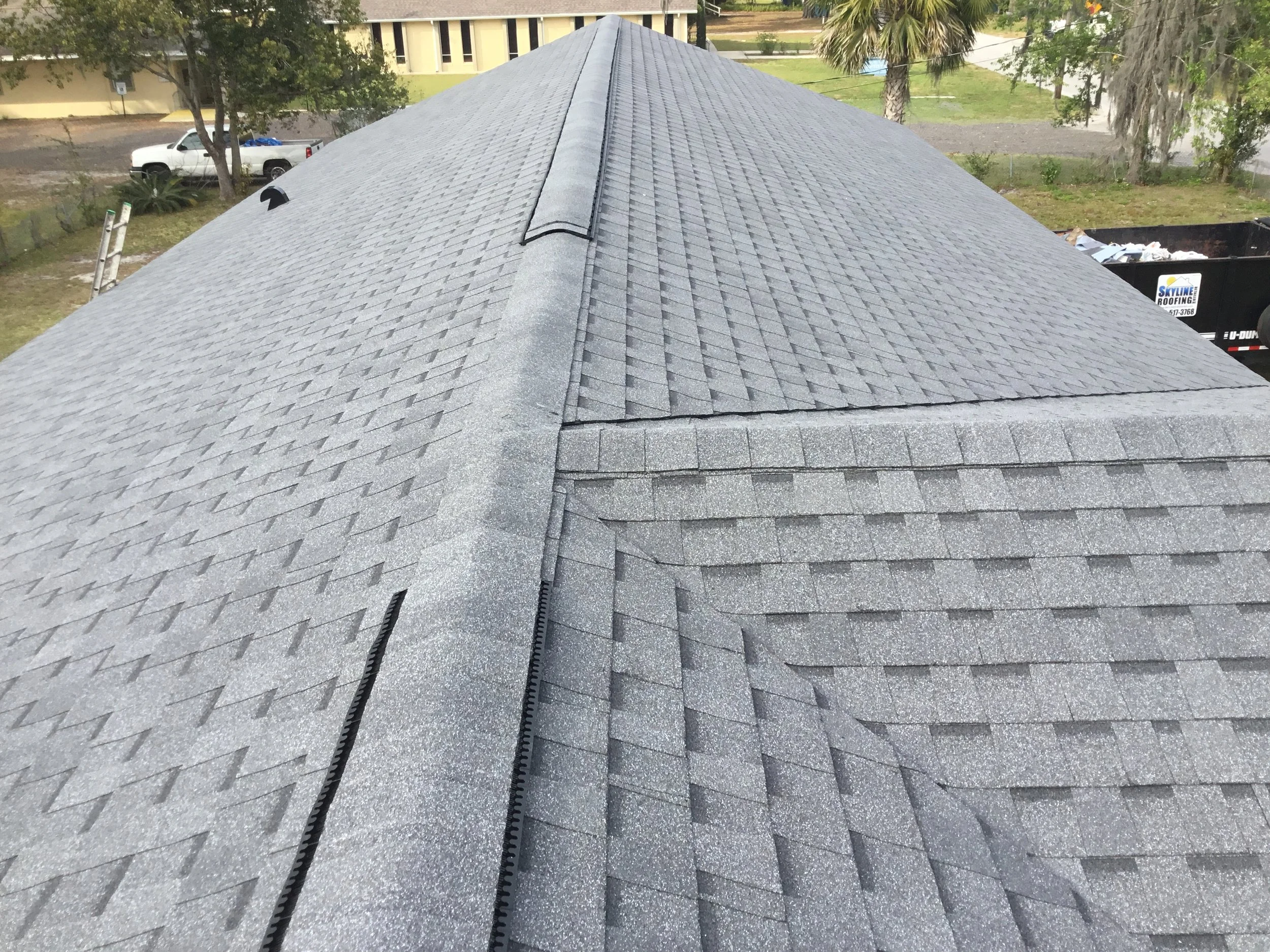 A new gray asphalt shingle roof with ridge cap and side flashing, with some ladders and trees in the background.
