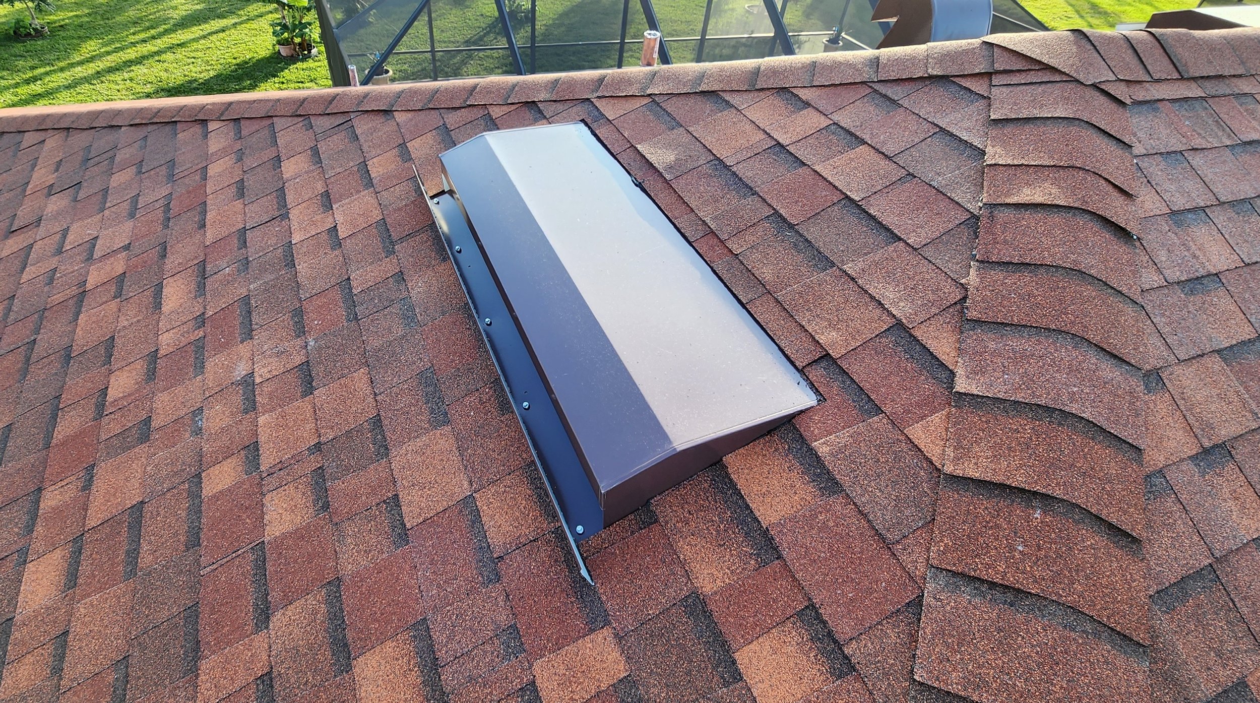 A metal skylight installed on a shingled roof with reddish-brown asphalt shingles, showing a scare with a field of grass and a glass greenhouse in the background.