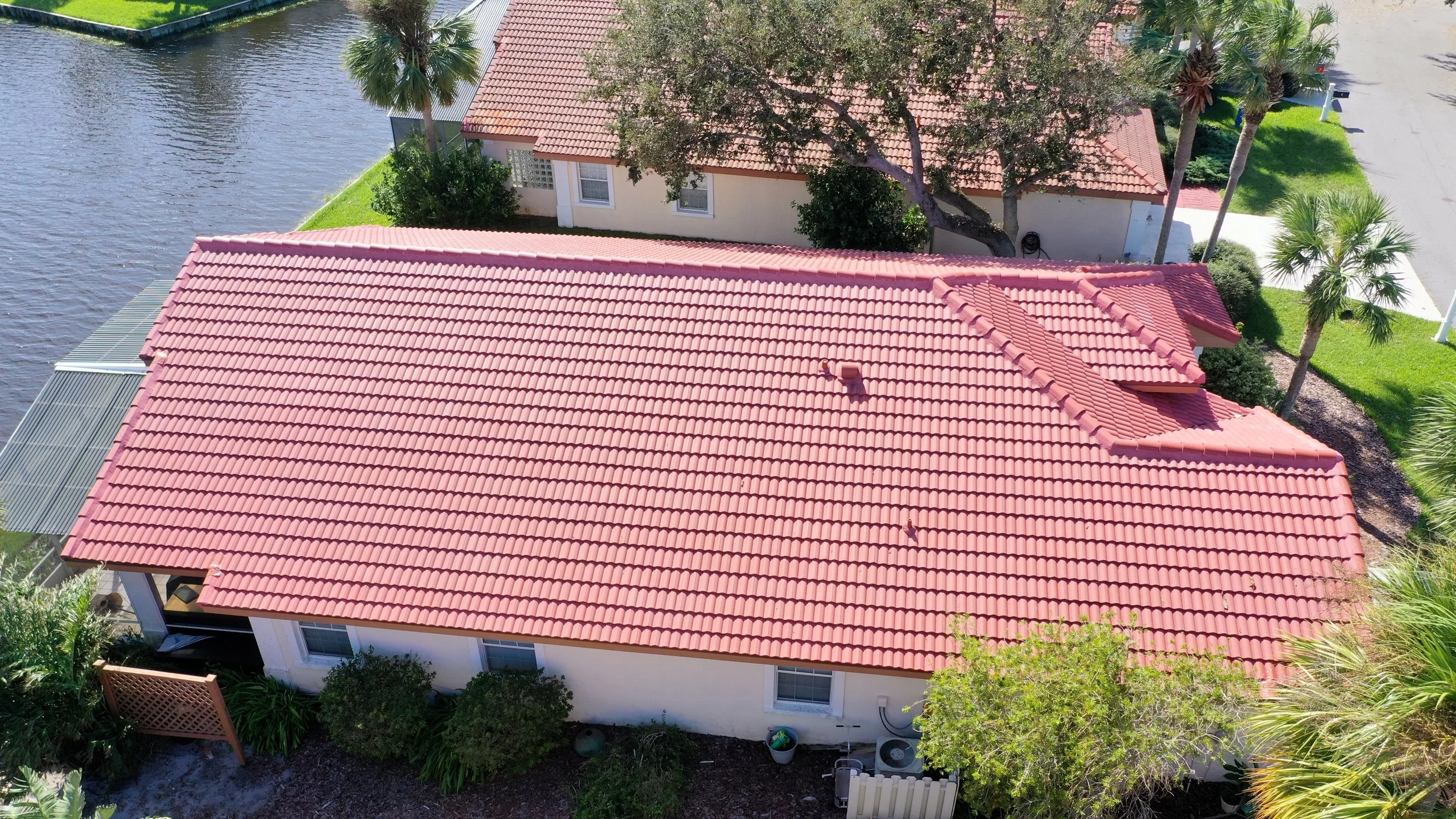 Aerial view of a residential house with a red tiled roof, surrounded by palm trees and green landscaping, next to a body of water.