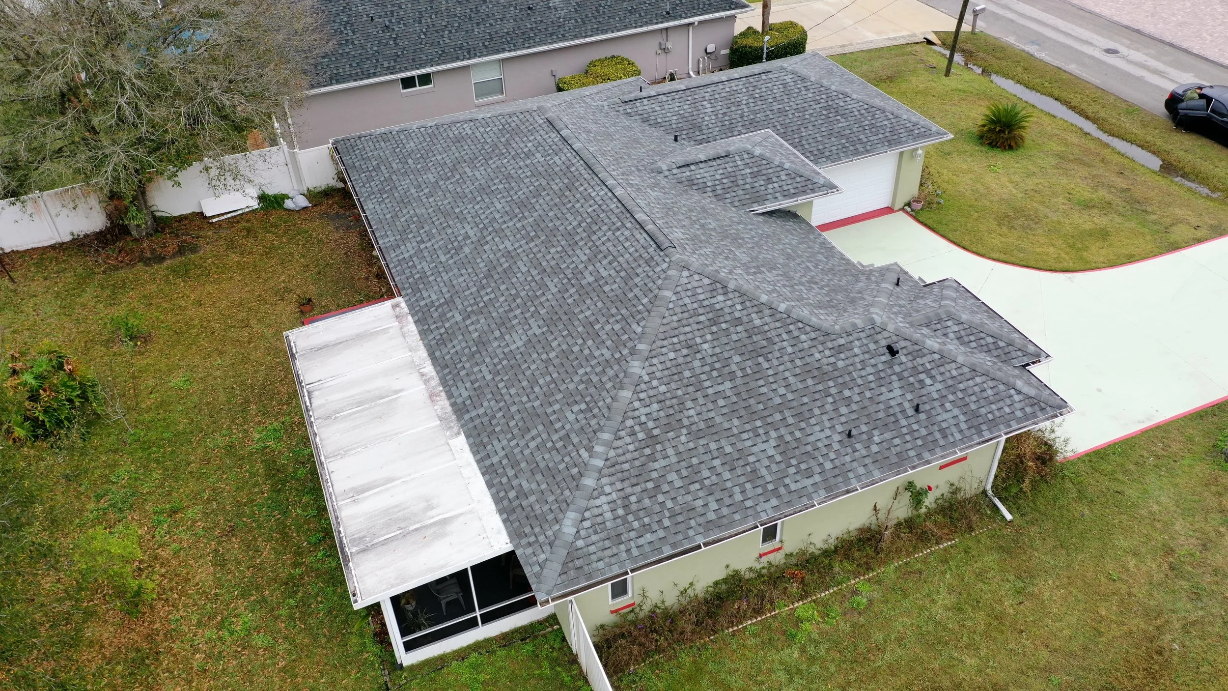 Aerial view of a house with a gray shingled roof, surrounded by a lawn, driveway, and backyard with a white fence and some trees.