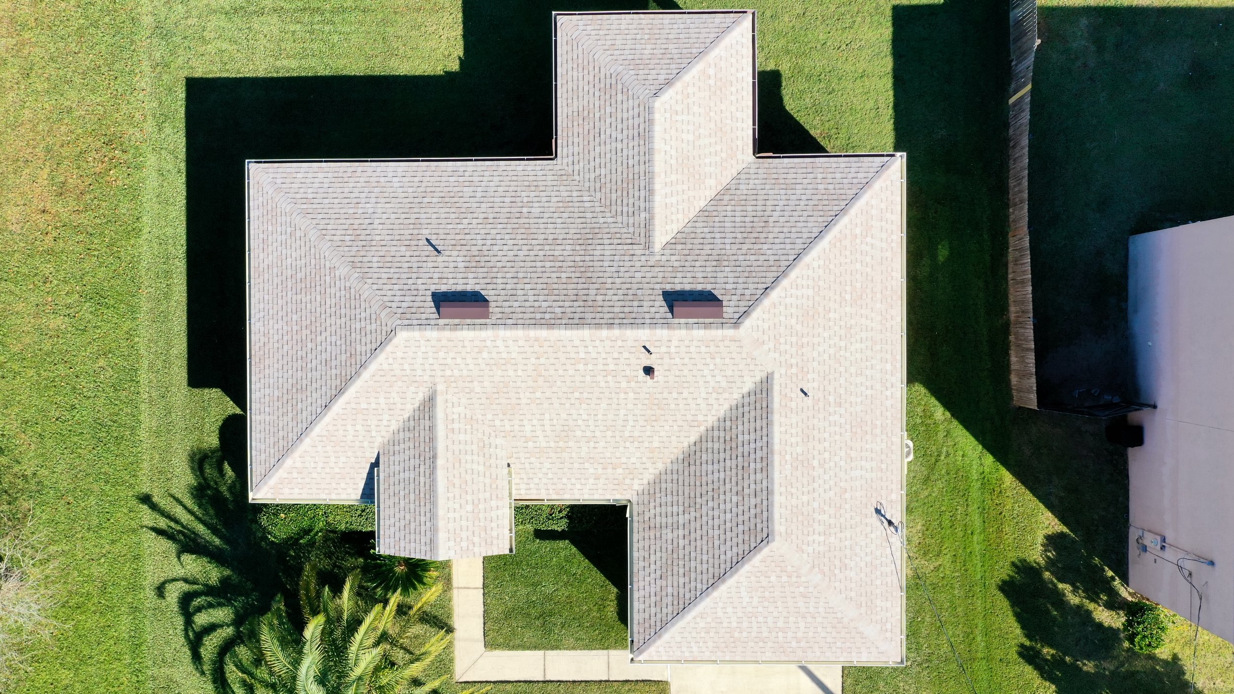 Top-down aerial view of a house with a light-colored shingled roof, surrounded by a green lawn and a sidewalk leading to the front entrance. Shadows of trees are visible on the grass.
