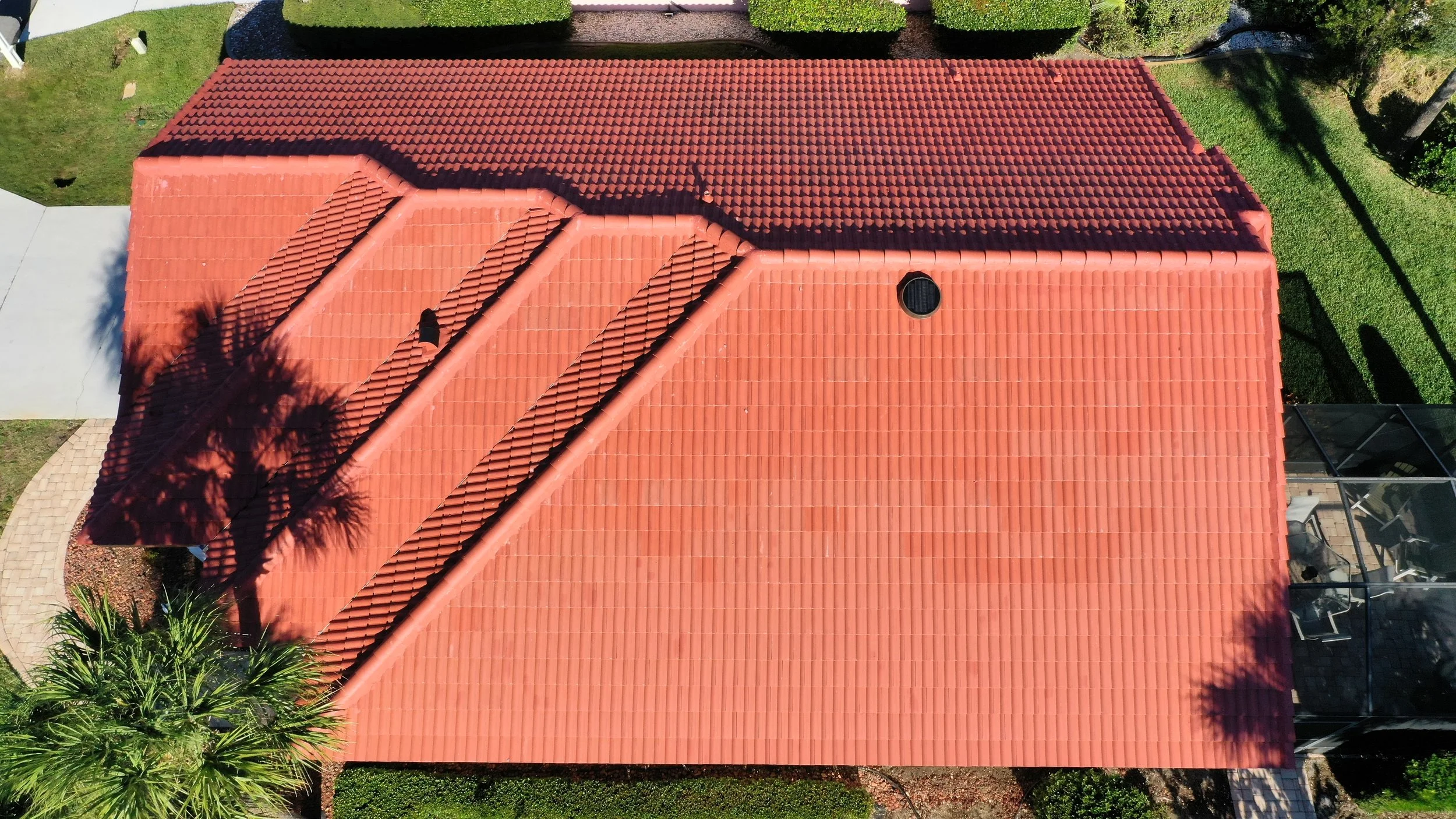 Aerial view of a house with a red tile roof, surrounded by green trees and lawn. There is a screened porch on the right side of the house and a paved walkway on the left.