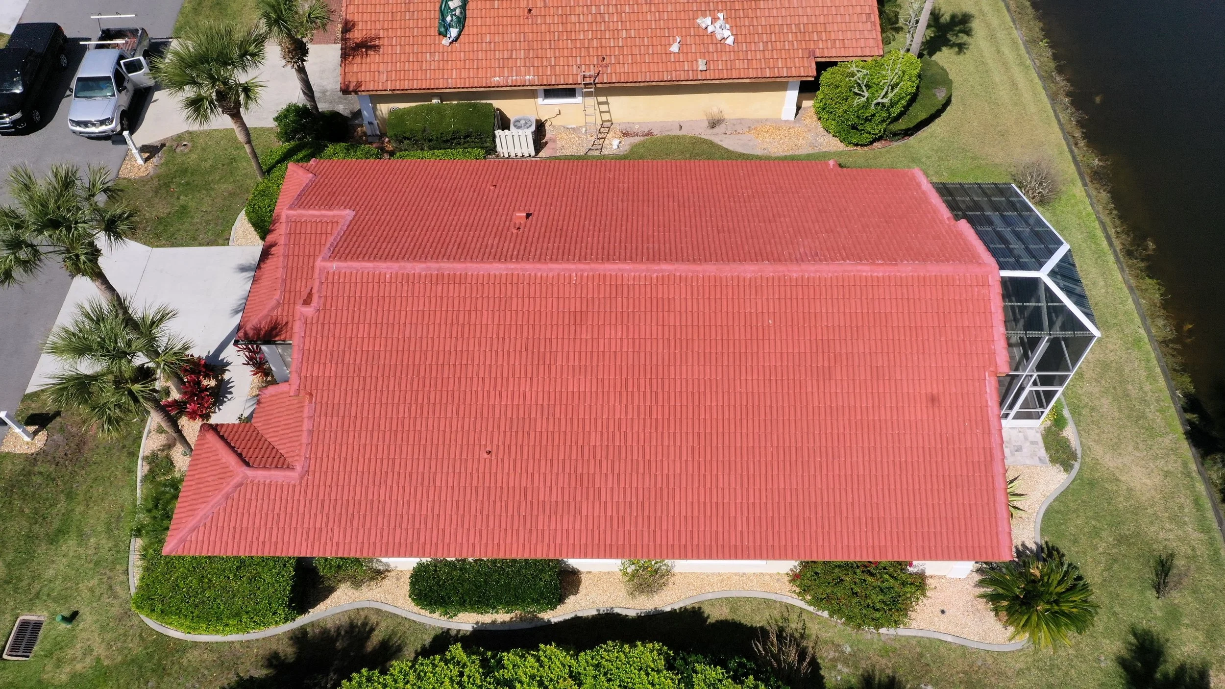 Aerial view of a house with a red tile roof and a screened porch, surrounded by palm trees and a lawn, adjacent to a waterway.