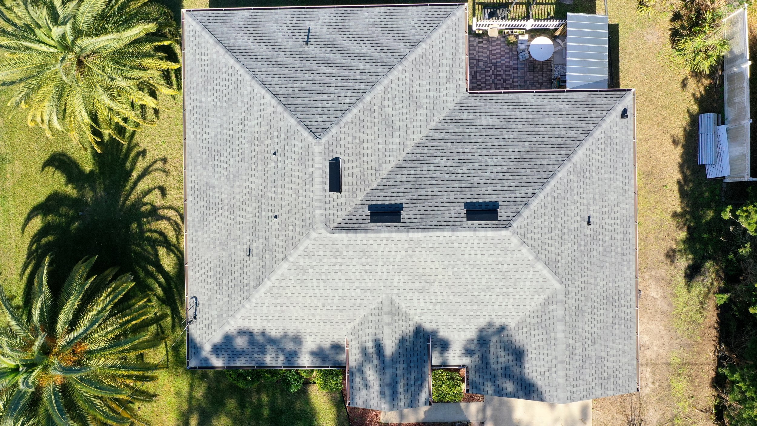 Aerial view of a house with a gray shingle roof, surrounded by green grass, palm trees, and some fenced yards.