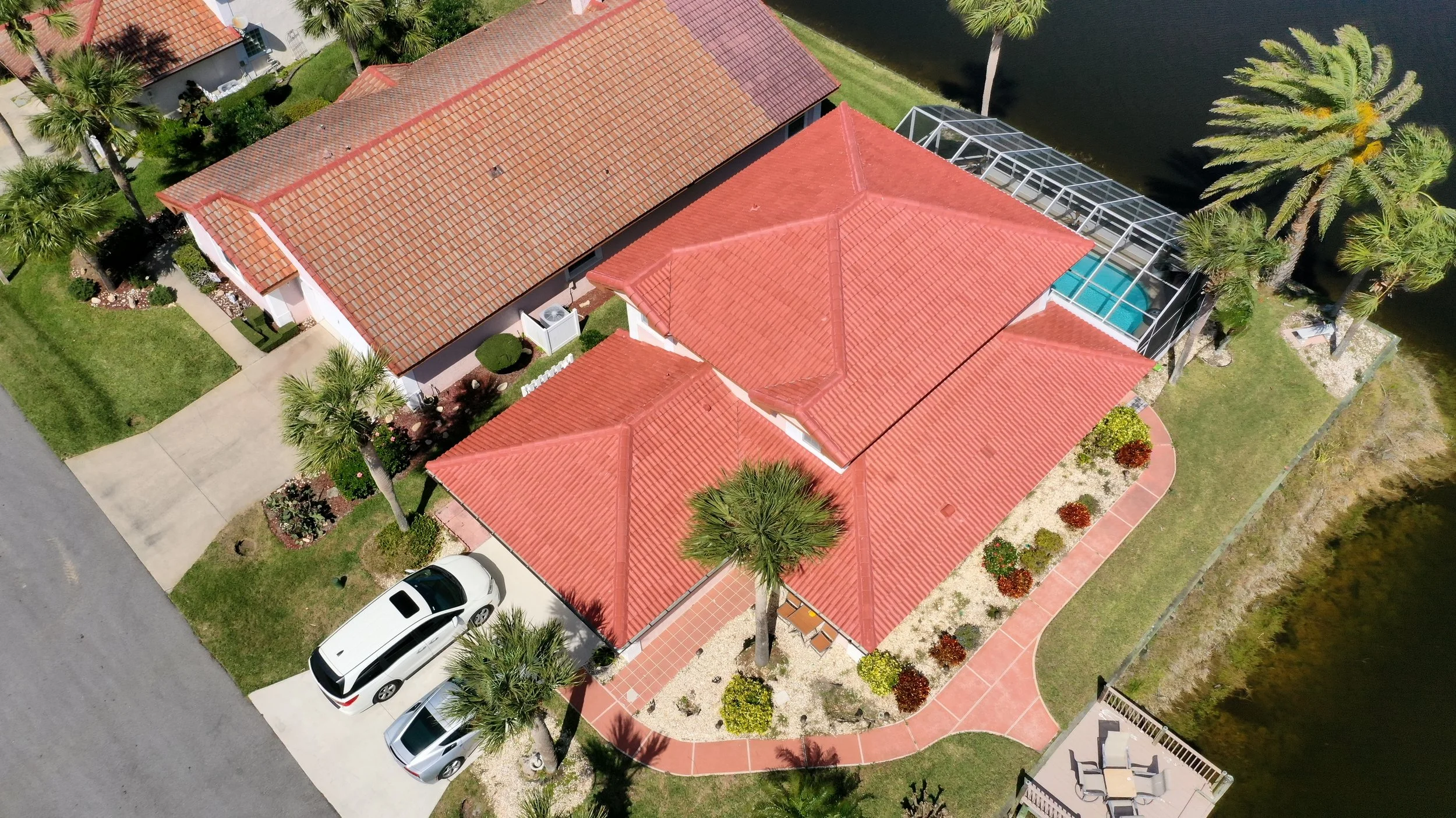 Aerial view of a residential house with red tile roof, landscaped yard, palm trees, a driveway with parked cars, and a screened-in swimming pool area near a water body.