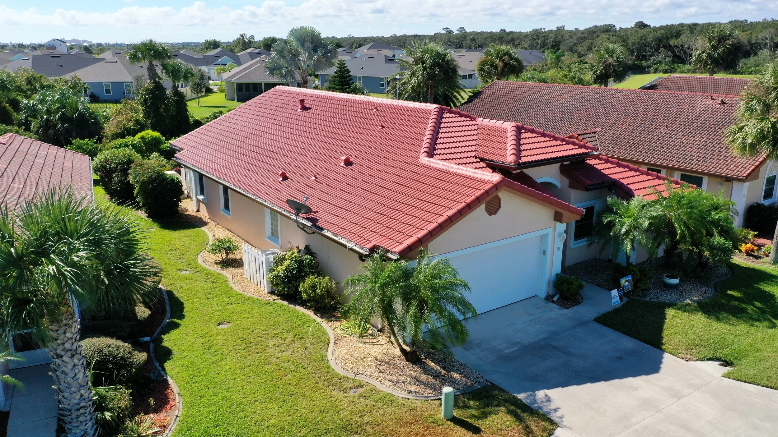 Aerial view of a residential neighborhood with houses featuring red-tile roofs, lush green lawns, and tropical trees, including palm trees, under a partly cloudy sky.