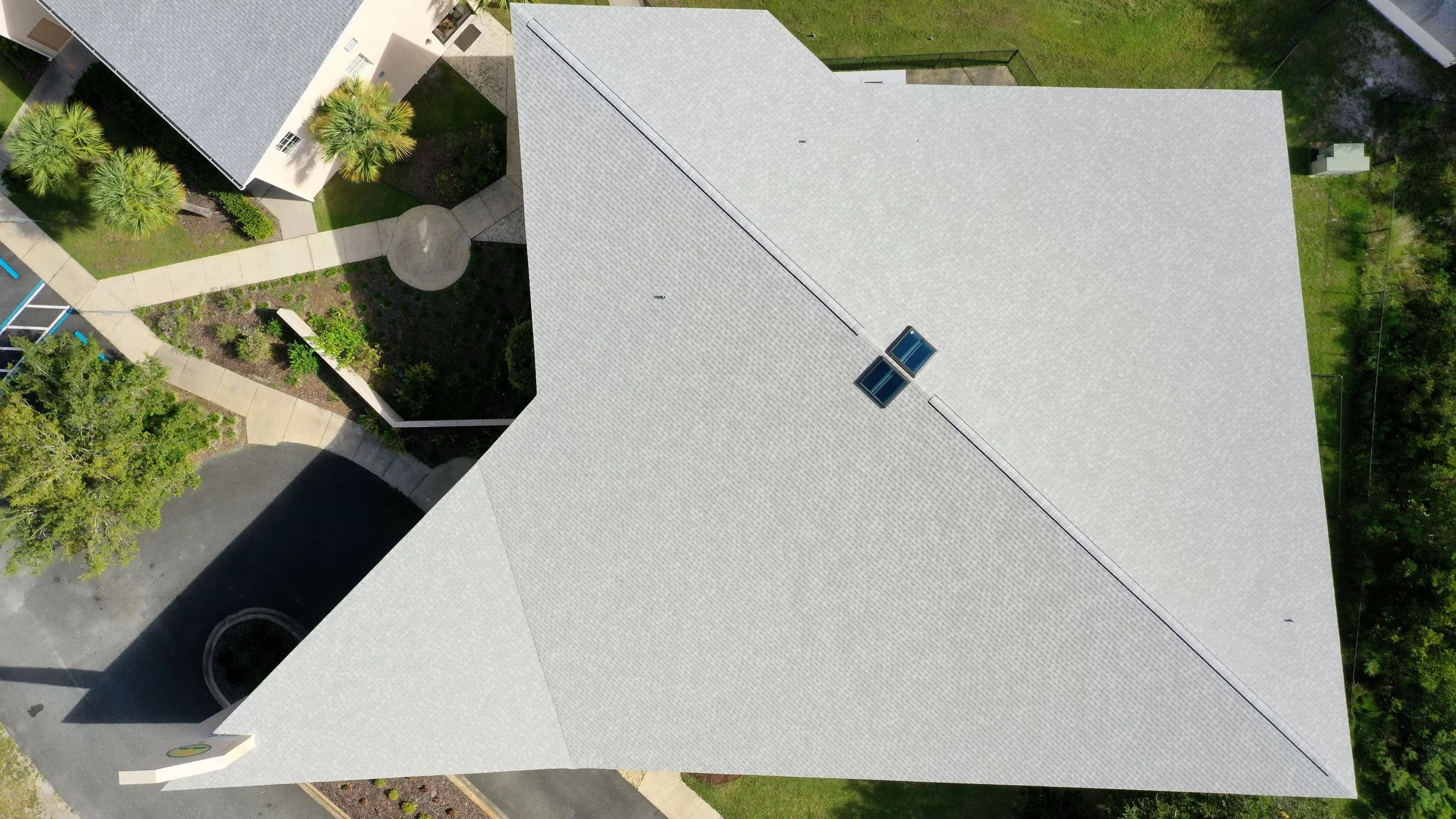 Aerial view of a residential building with a gray shingle roof, two skylights, surrounded by trees, a driveway, and a landscape garden.