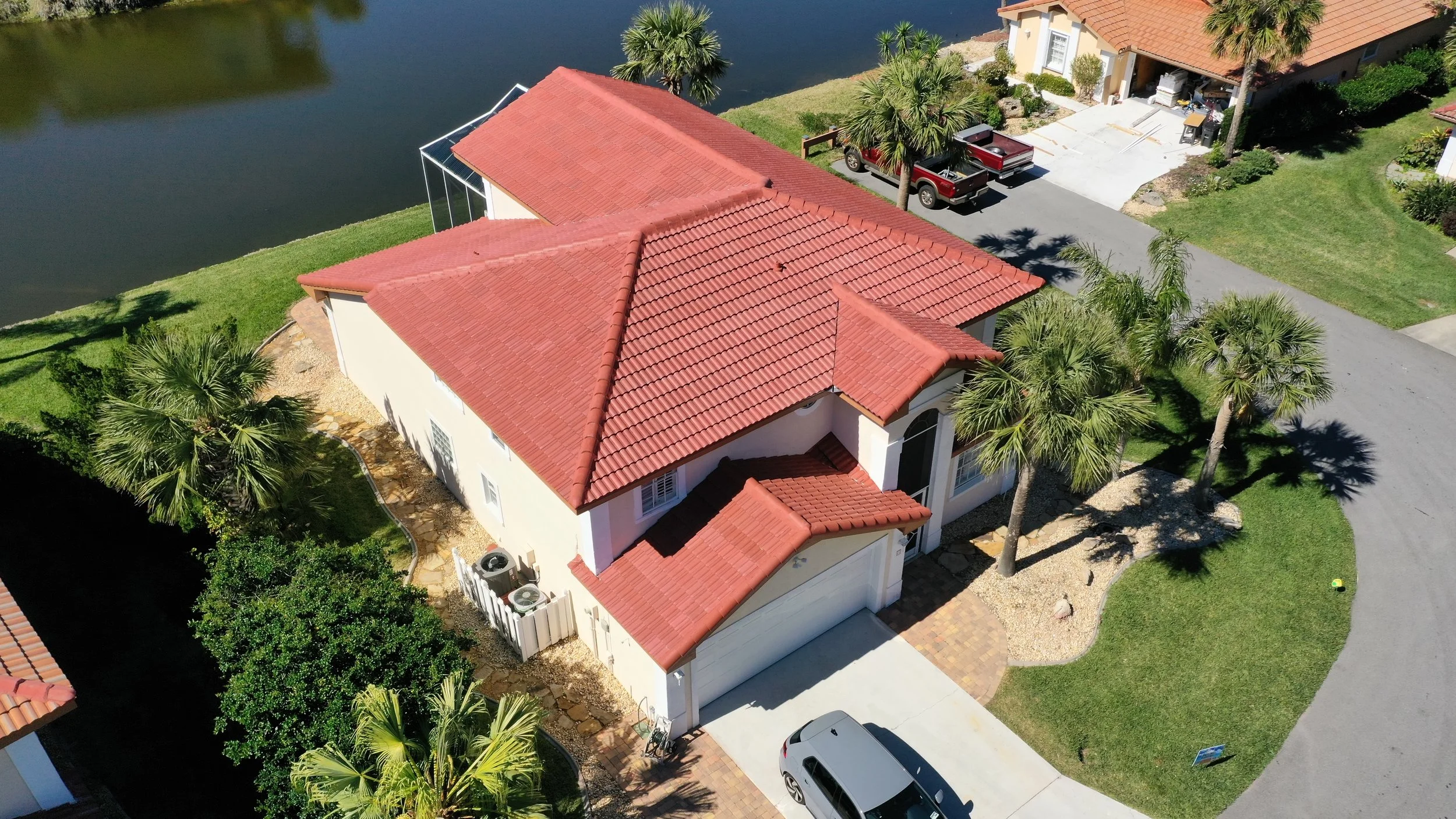 Aerial view of a house with a red tiled roof, surrounded by palm trees and green lawn, near a body of water. There are parked cars on the driveway and a boat lift in the water.