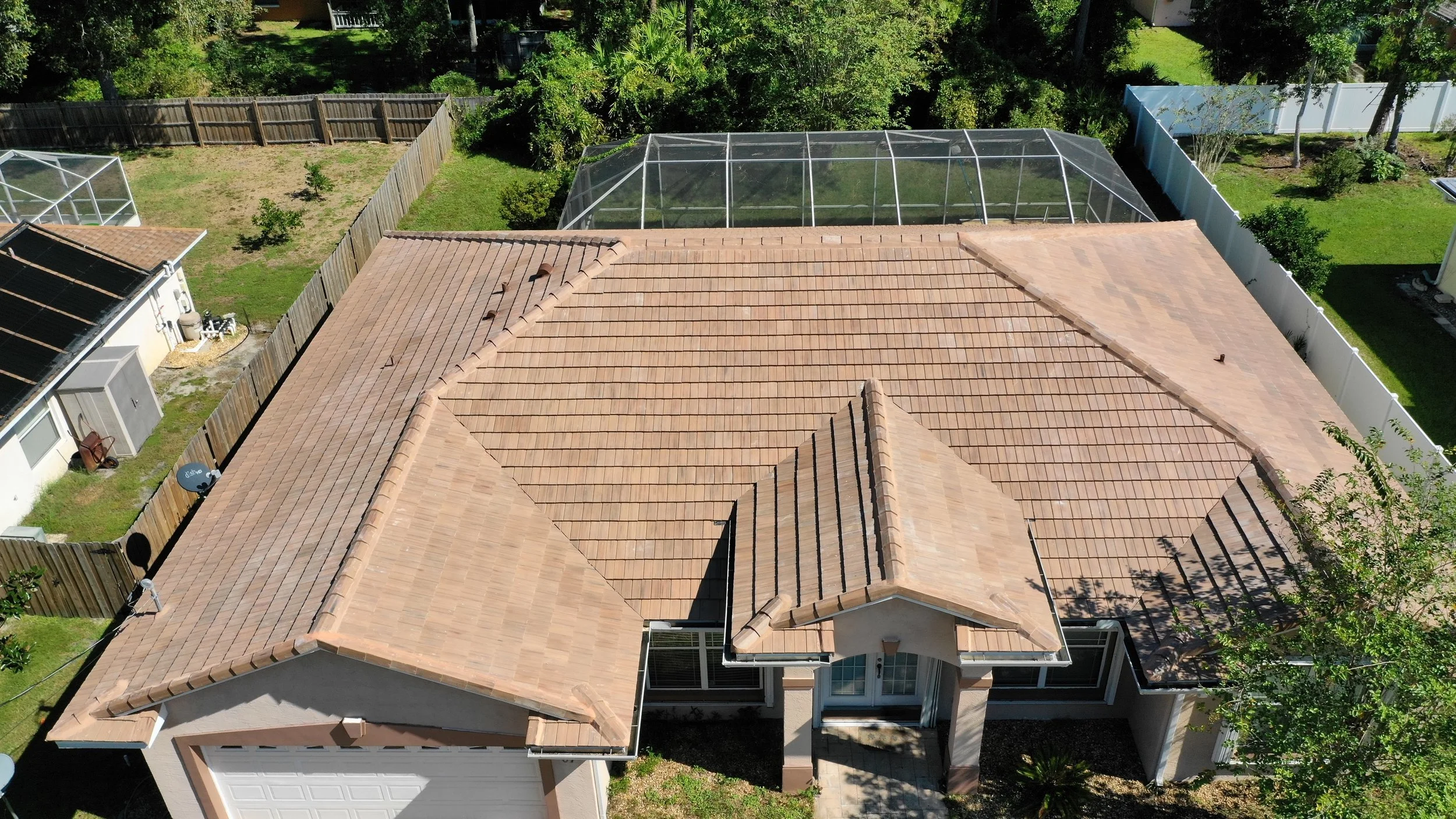 Aerial view of a house with a tan tiled roof, surrounded by fences, with neighboring yards featuring trees, a greenhouse, and a satellite dish.