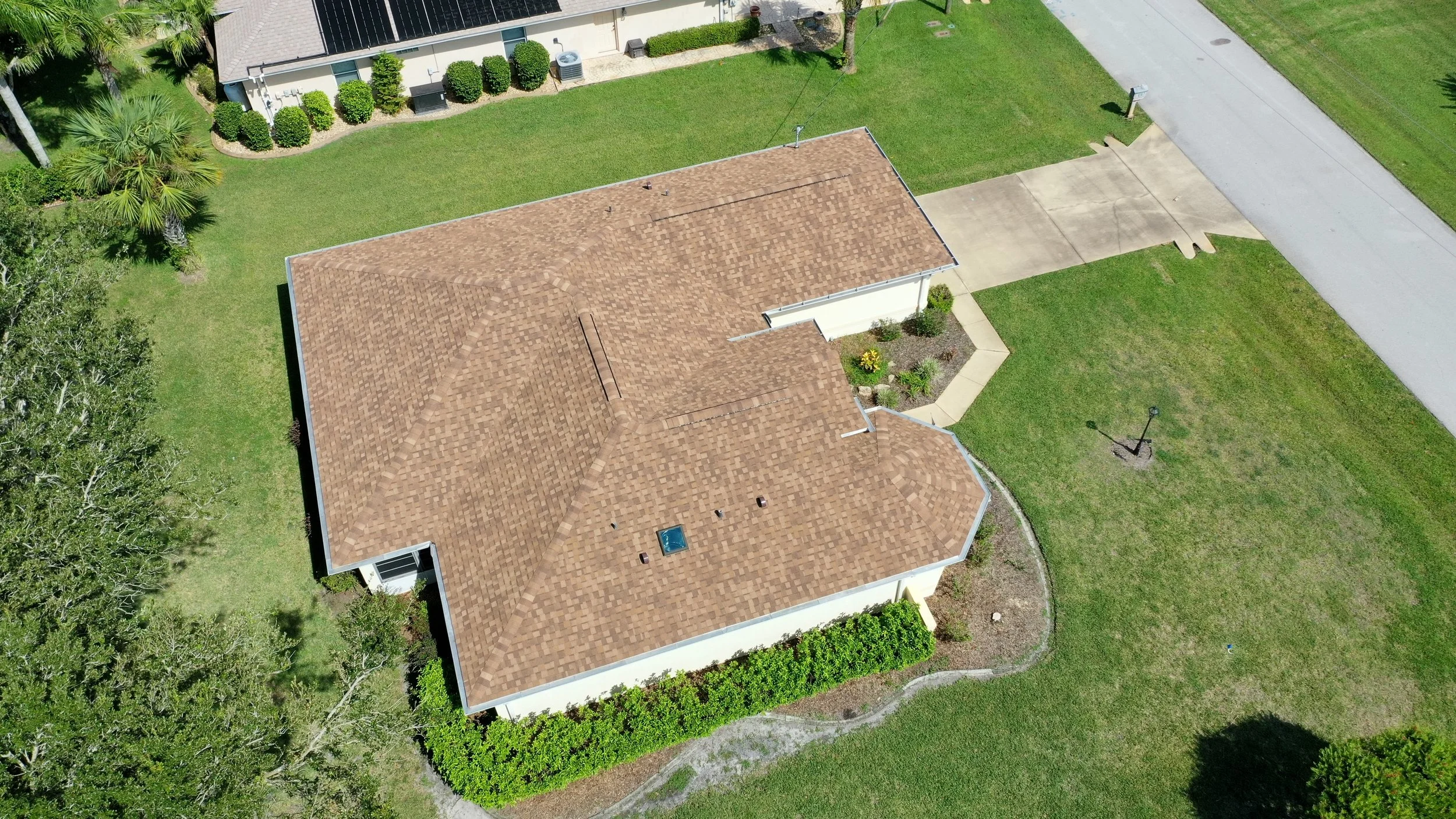 Aerial view of a house with tan shingle roof, surrounded by green lawn, bushes, trees, and a concrete driveway.