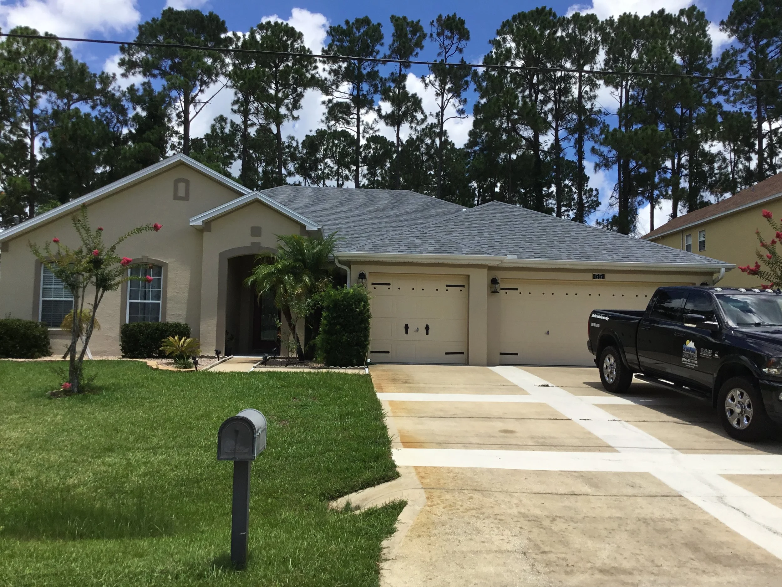A suburban house with a beige exterior, gray shingle roof, and two garage doors. The front yard has green grass, small shrubs, and a flowering tree. A black pickup truck is parked in the driveway, and there is a mailbox in the foreground. Tall trees 