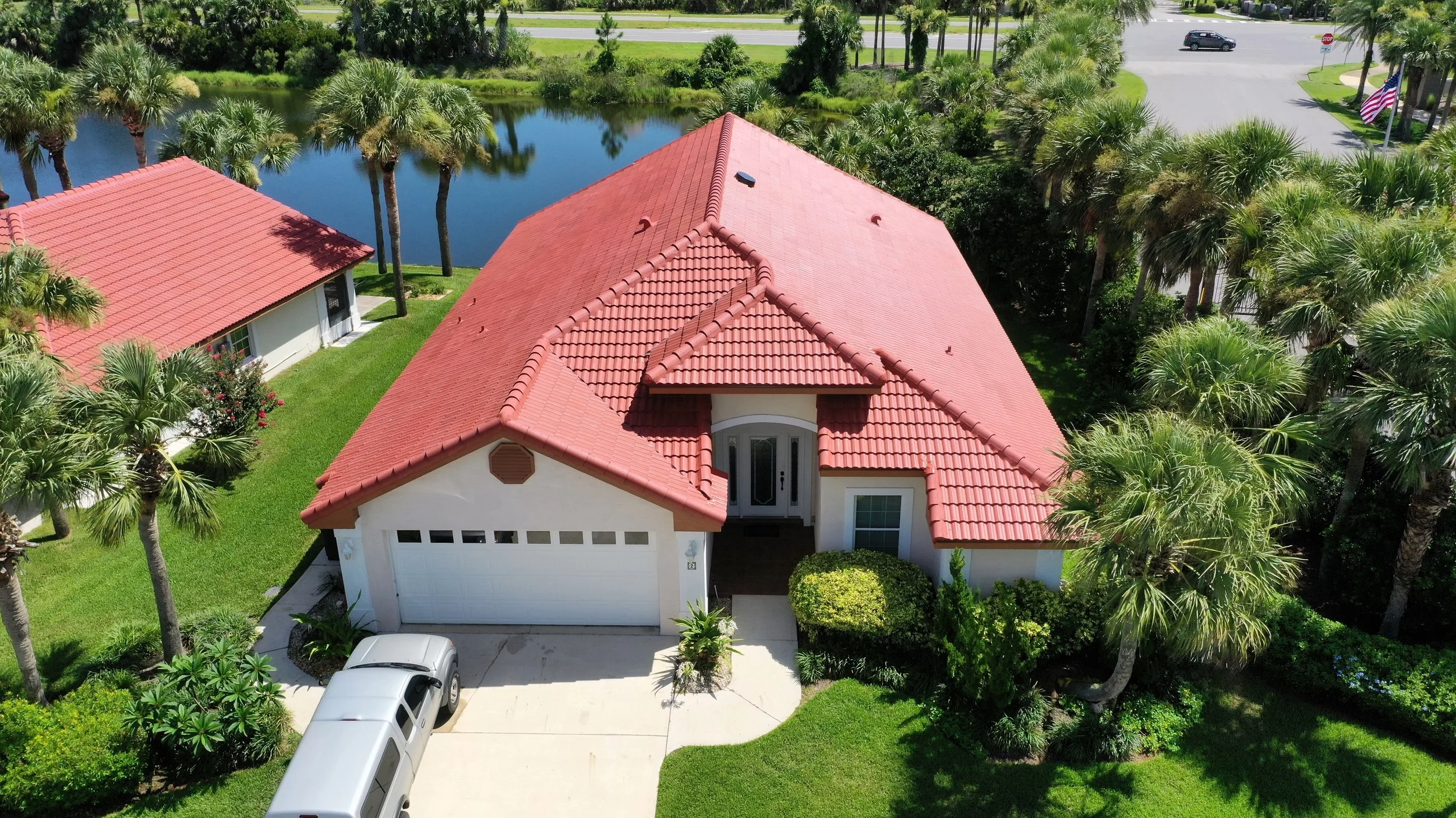 Aerial view of a house with a red-tile roof, white walls, a two-car garage, and a driveway with a silver vehicle parked on it. The house is surrounded by green grass, palm trees, and shrubs. There is a body of water and a road with cars in the backgr