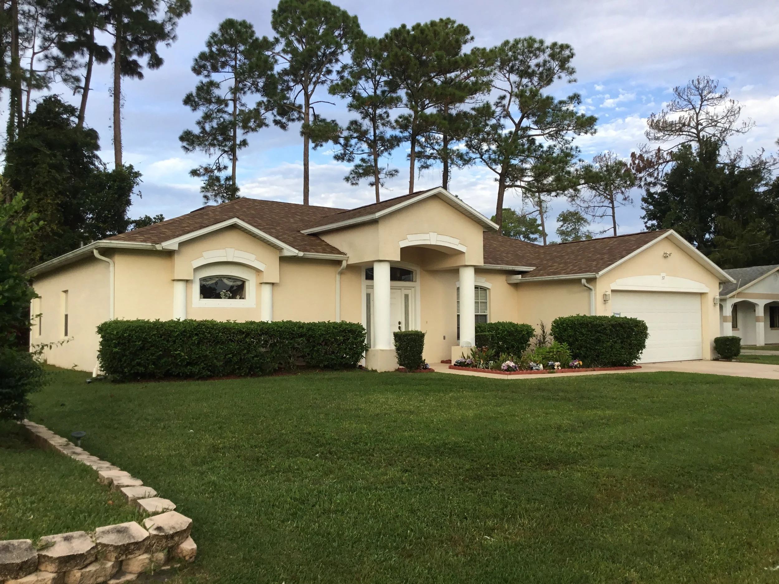 A beige single-story house with a brown shingle roof, white columns at the entrance, a two-car garage, landscaped front yard, green bushes, flower bed, and tall pine trees in the background.