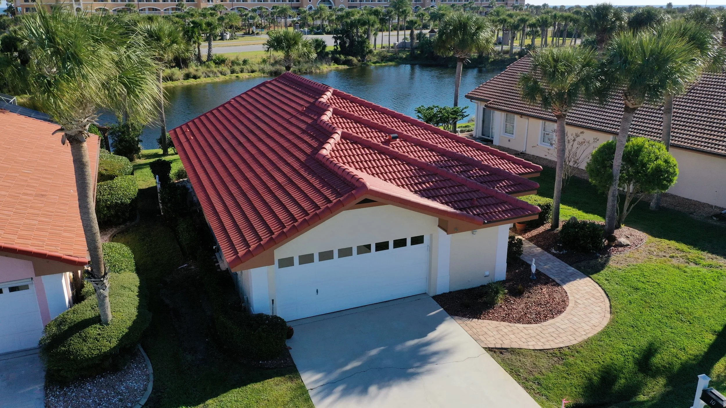 Aerial view of a suburban house with a red tile roof, surrounded by green lawns, trees, and bushes, with a body of water nearby and other houses in the neighborhood.