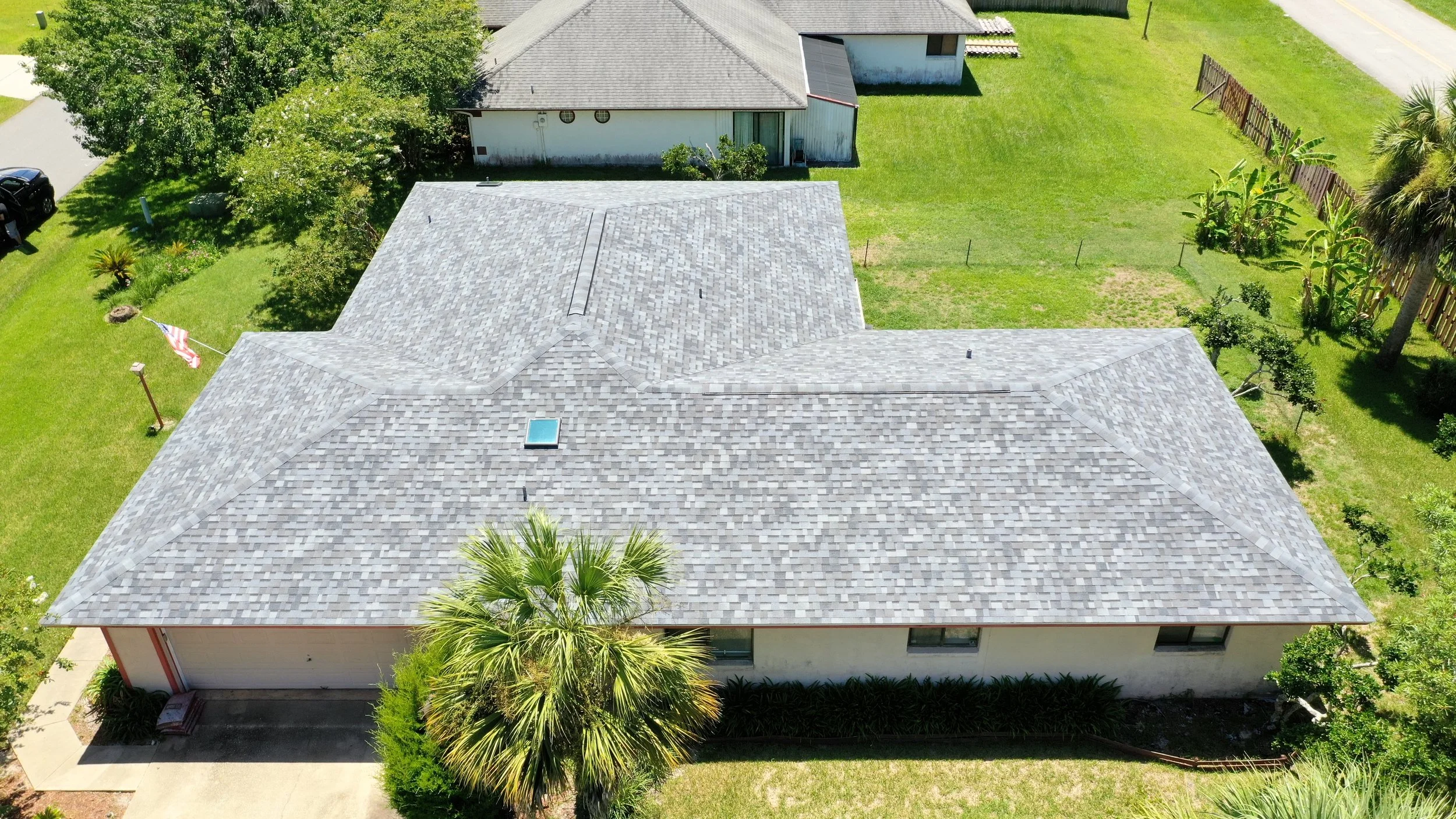 Aerial view of a house with a gray shingle roof, surrounded by a green lawn, trees, and a wooden fence in a suburban neighborhood.