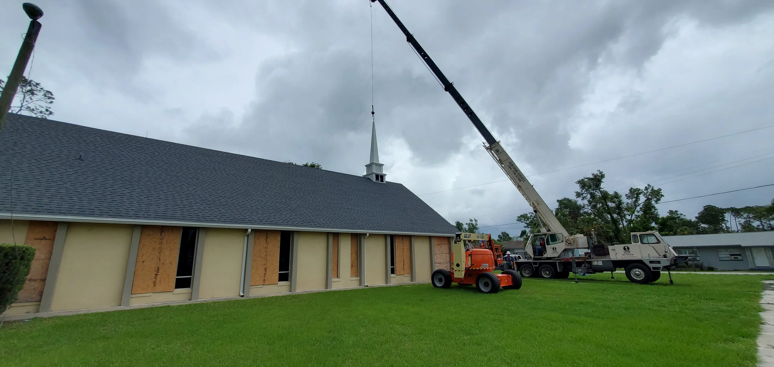 Crane lifts a church steeple during construction or repair work on a church building with boarded-up windows and a sloped roof, on a cloudy day.