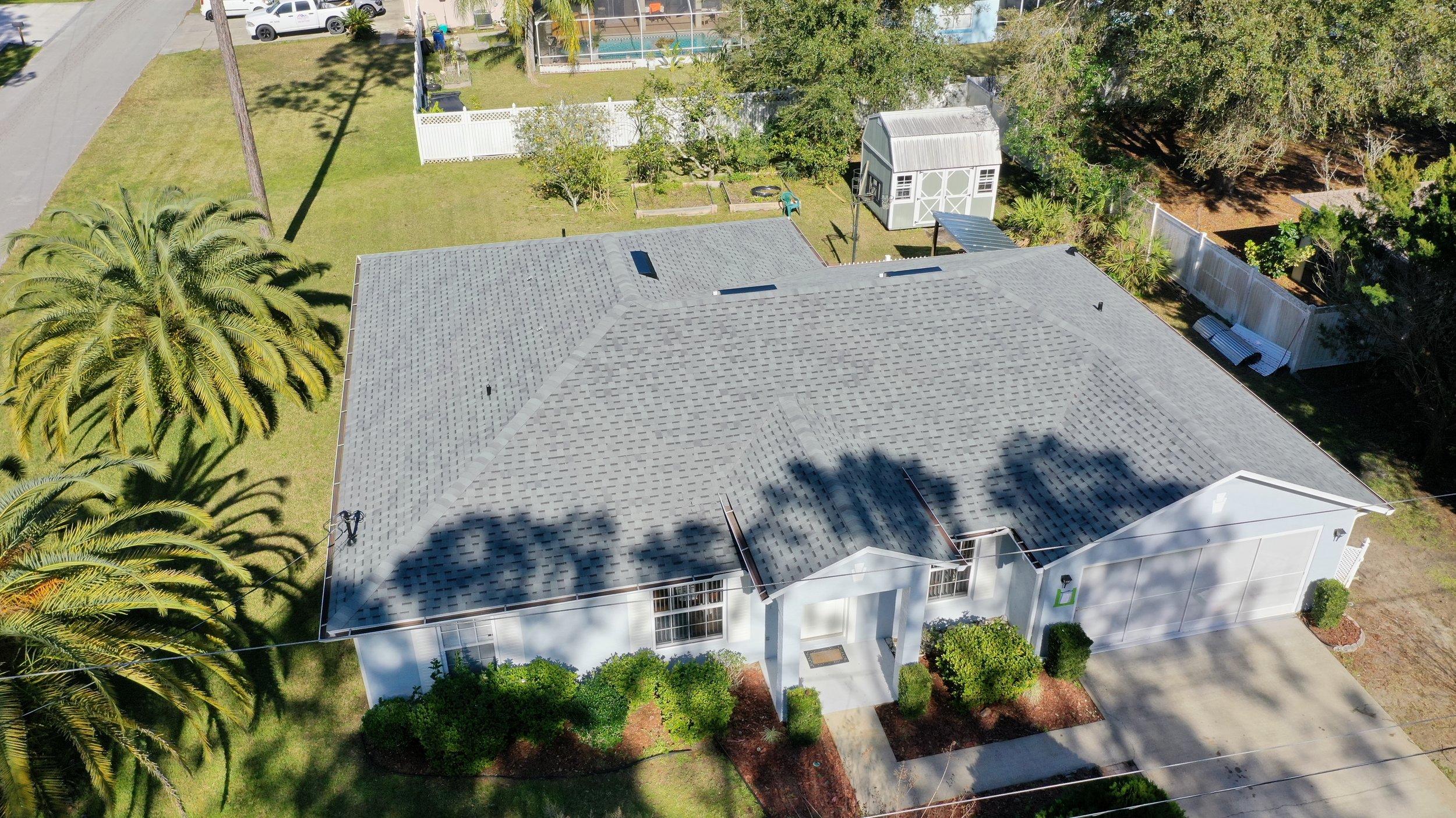 Aerial view of a house with a grey shingle roof, white exterior walls, surrounding green lawn, palm trees, and a driveway leading to a garage. There are shadows cast on the roof and yard, and neighboring properties are visible in the background.