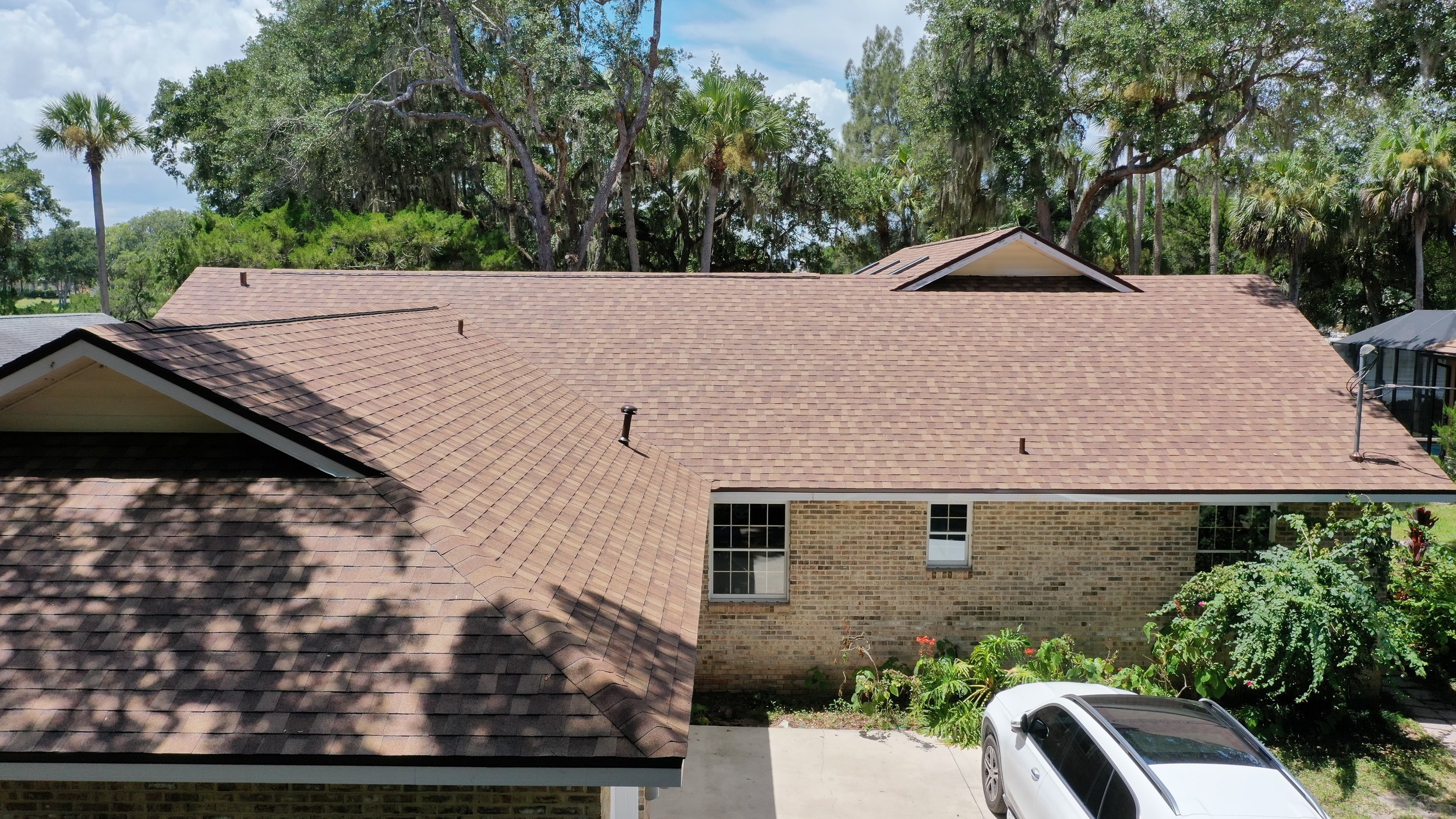 View of rooftops of two houses, one with brown shingles and one with gray shingles, surrounded by trees including palms and Oaks, with a white car parked in a driveway in front of a brick house.