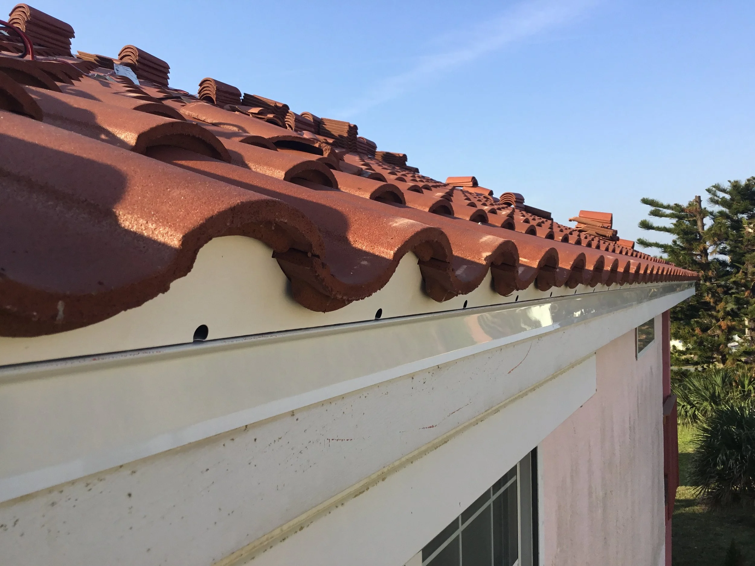 Close-up of a house roof with red clay tiles and a white gutter, with a clear blue sky and some trees in the background.
