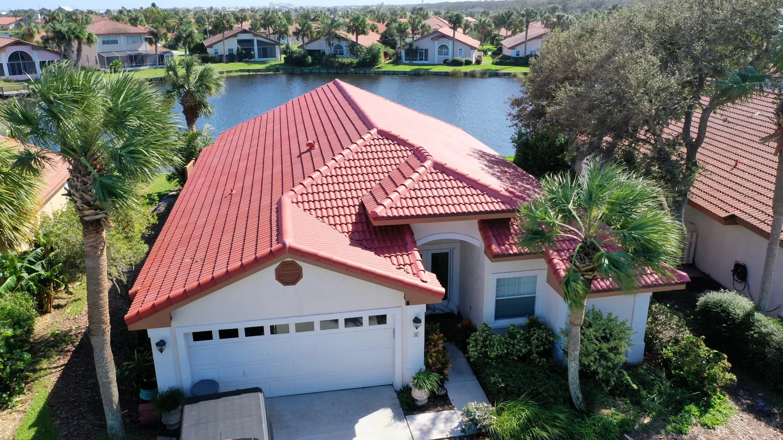 A house with a red tiled roof surrounded by palm trees and greenery. The house is located near a pond with similar houses in the background.