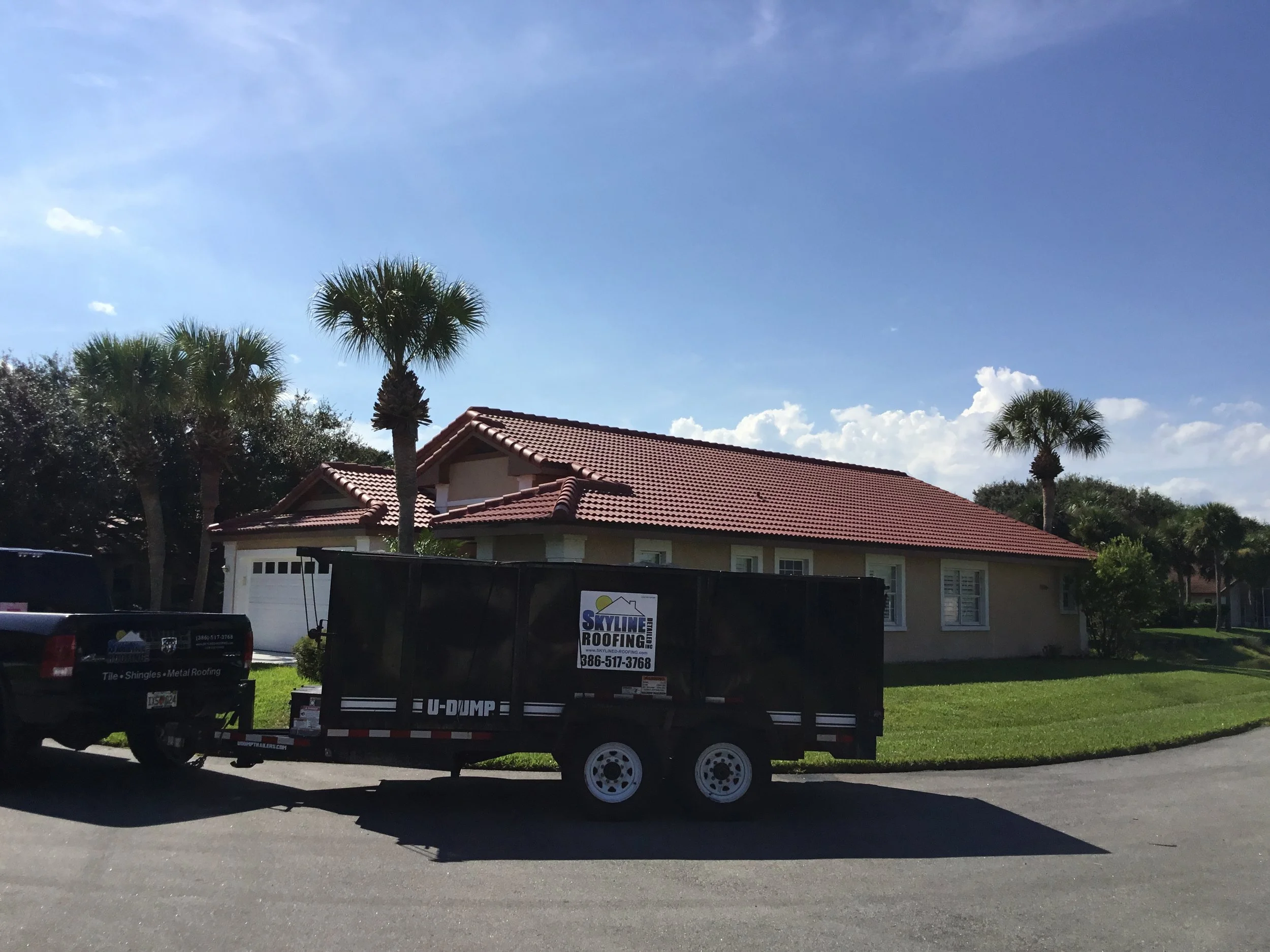 House with a red-tiled roof, white exterior walls, and palm trees in the front yard, parked on a paved driveway with a black trailer attached to a pickup truck.
