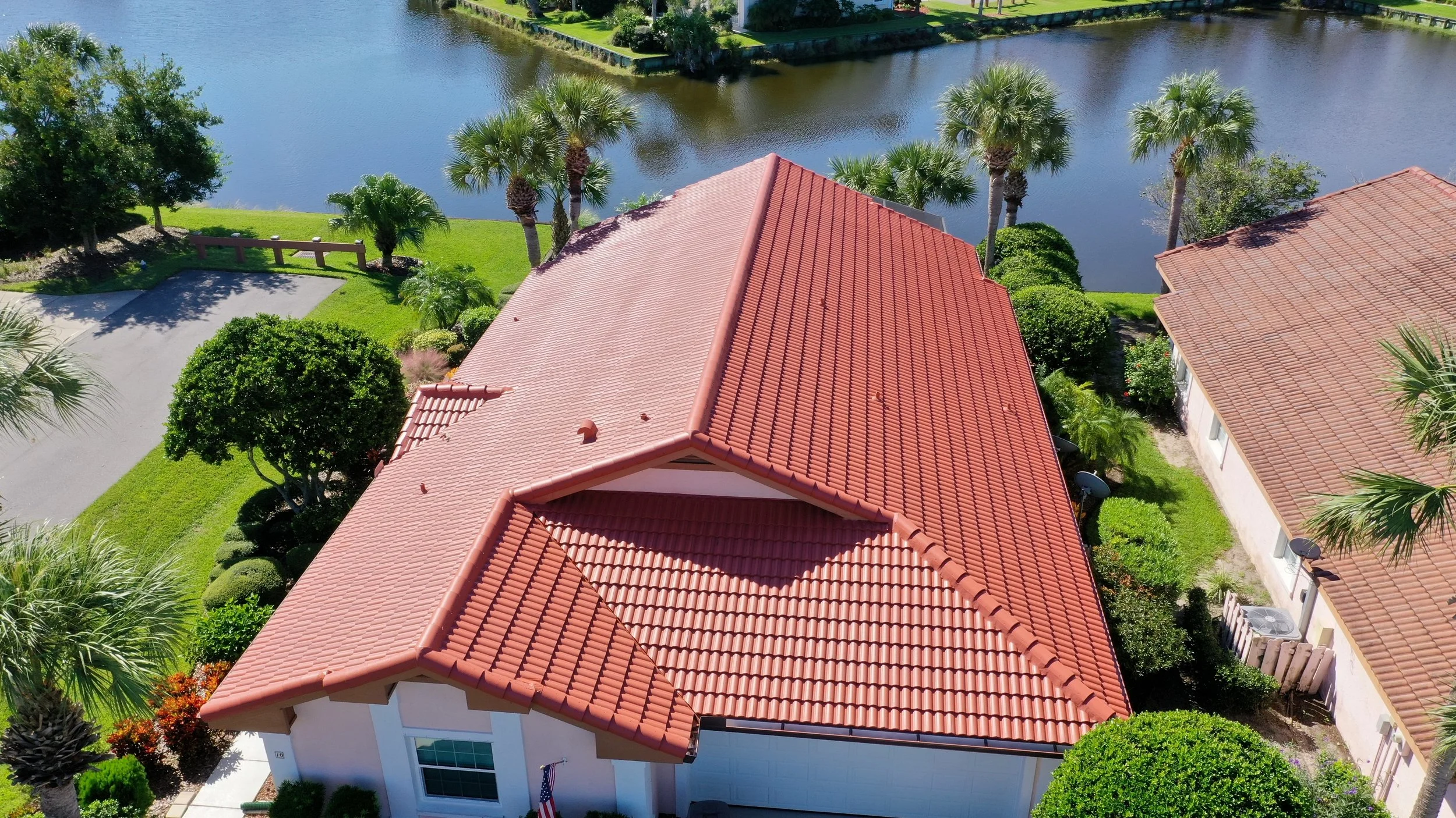 Aerial view of a house with a red tiled roof, surrounded by lush green trees, including palm trees, and near a body of water in a neighborhood.