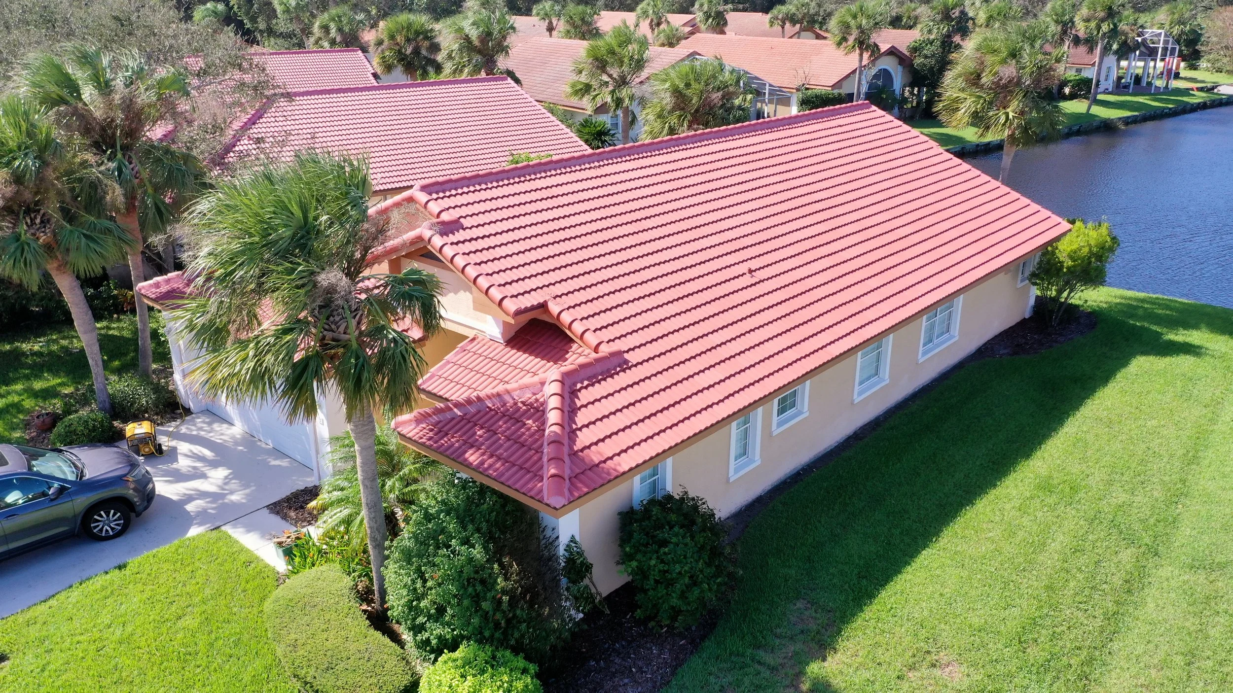 Aerial view of a suburban house with a red-tiled roof, surrounded by green lawn, palm trees, and a pond nearby.