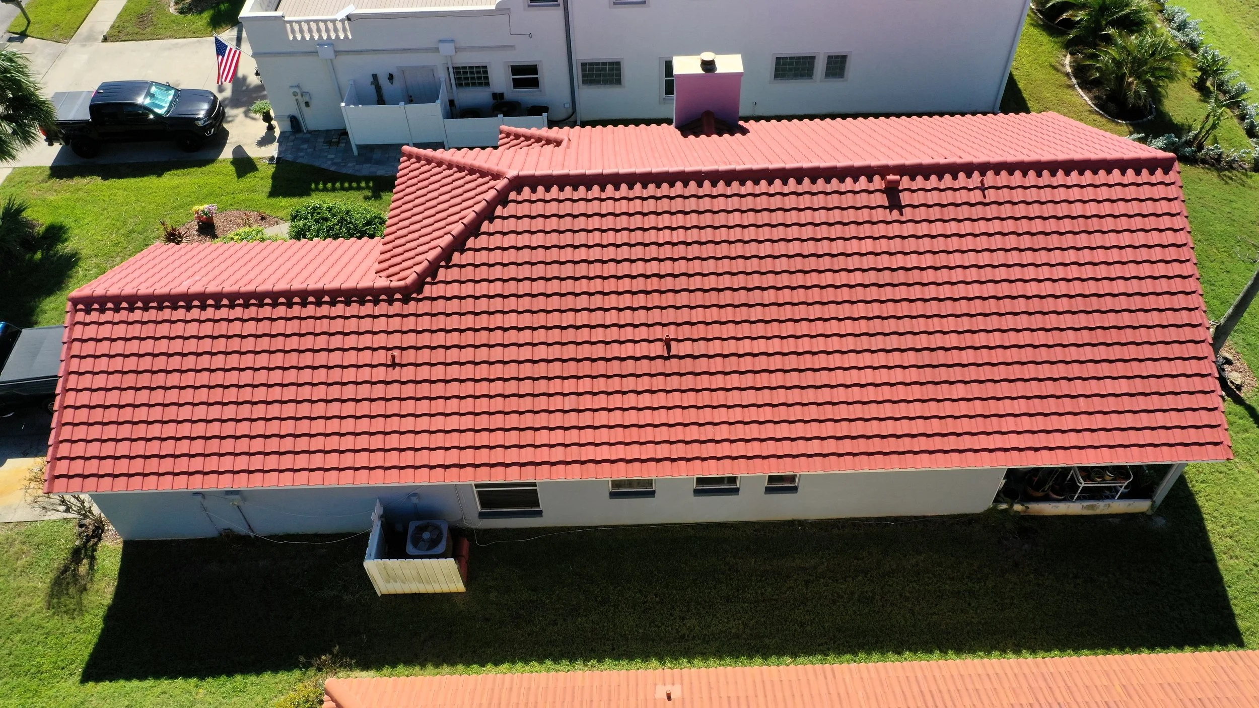 Aerial view of a house with a red tile roof, white exterior walls, and three small windows. The house has a small backyard with grass, a small garden with plants, and a patio. Nearby, there is a neighboring house with a white exterior, a parking area