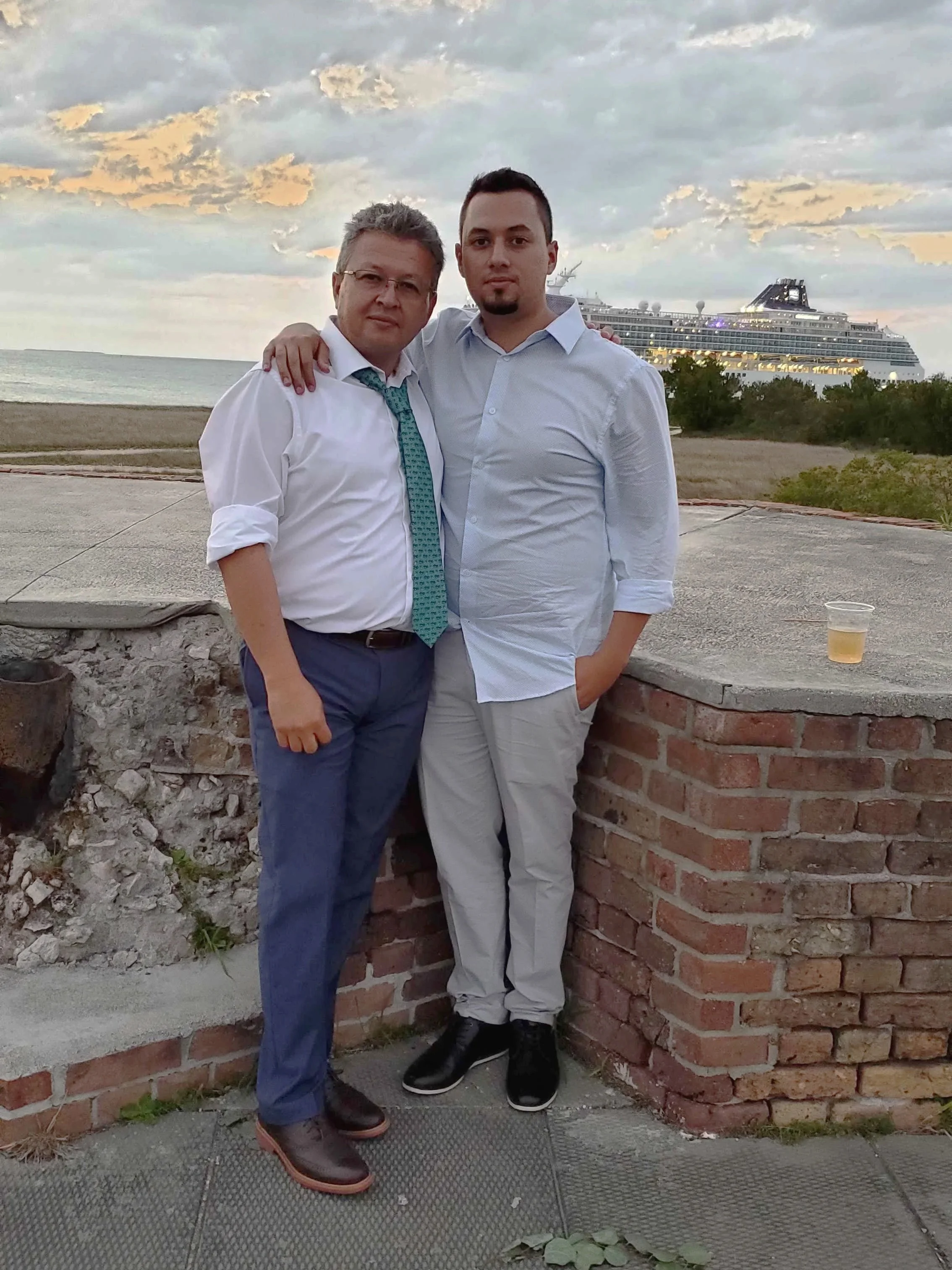 Two men standing close together with their arms around each other, posing outdoors near a brick wall with a cruise ship in the background during sunset.