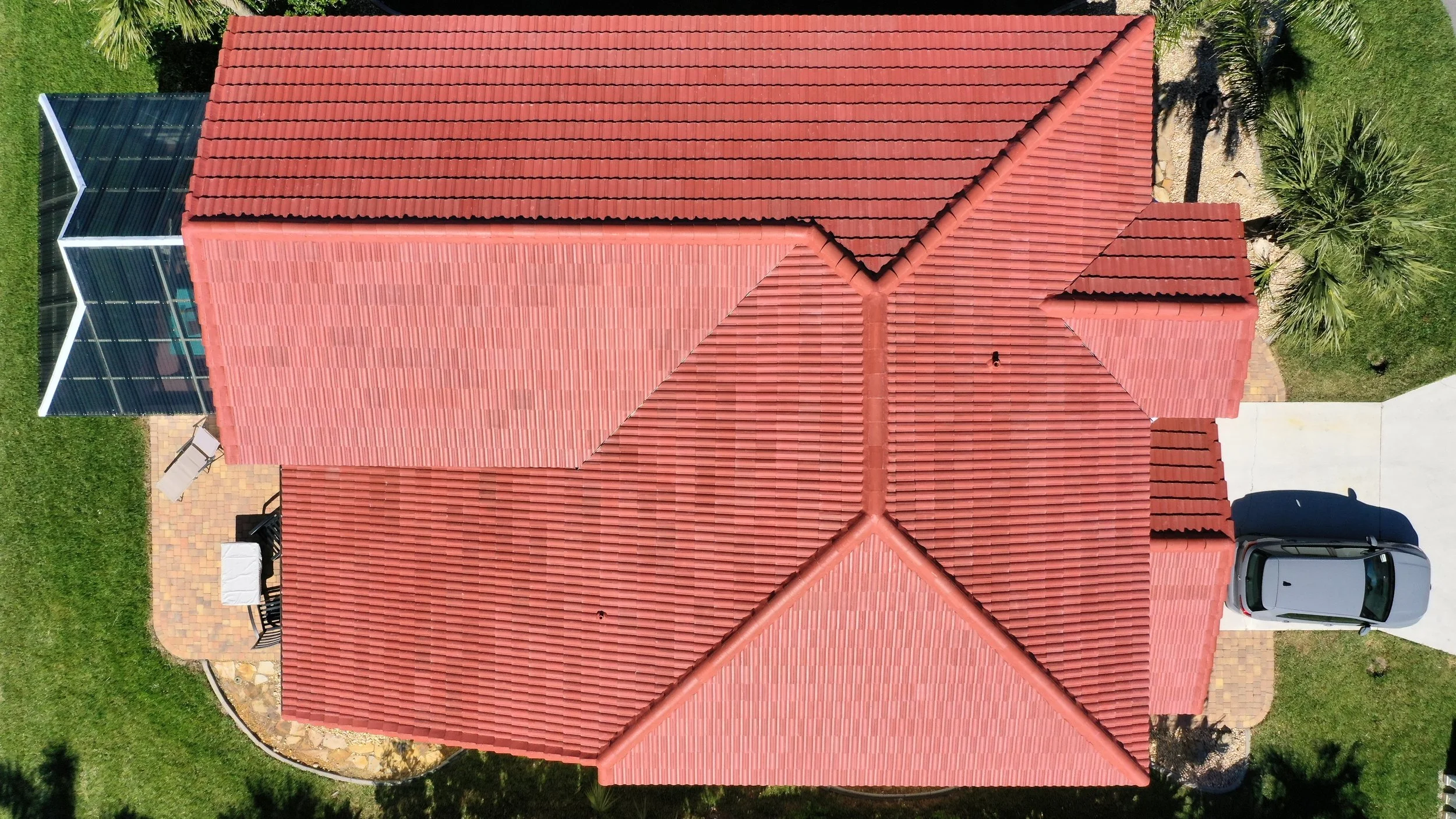 Top-down aerial view of a house with red tile roof, driveway with parked car, green lawn, palm trees, and a patio area with outdoor furniture.