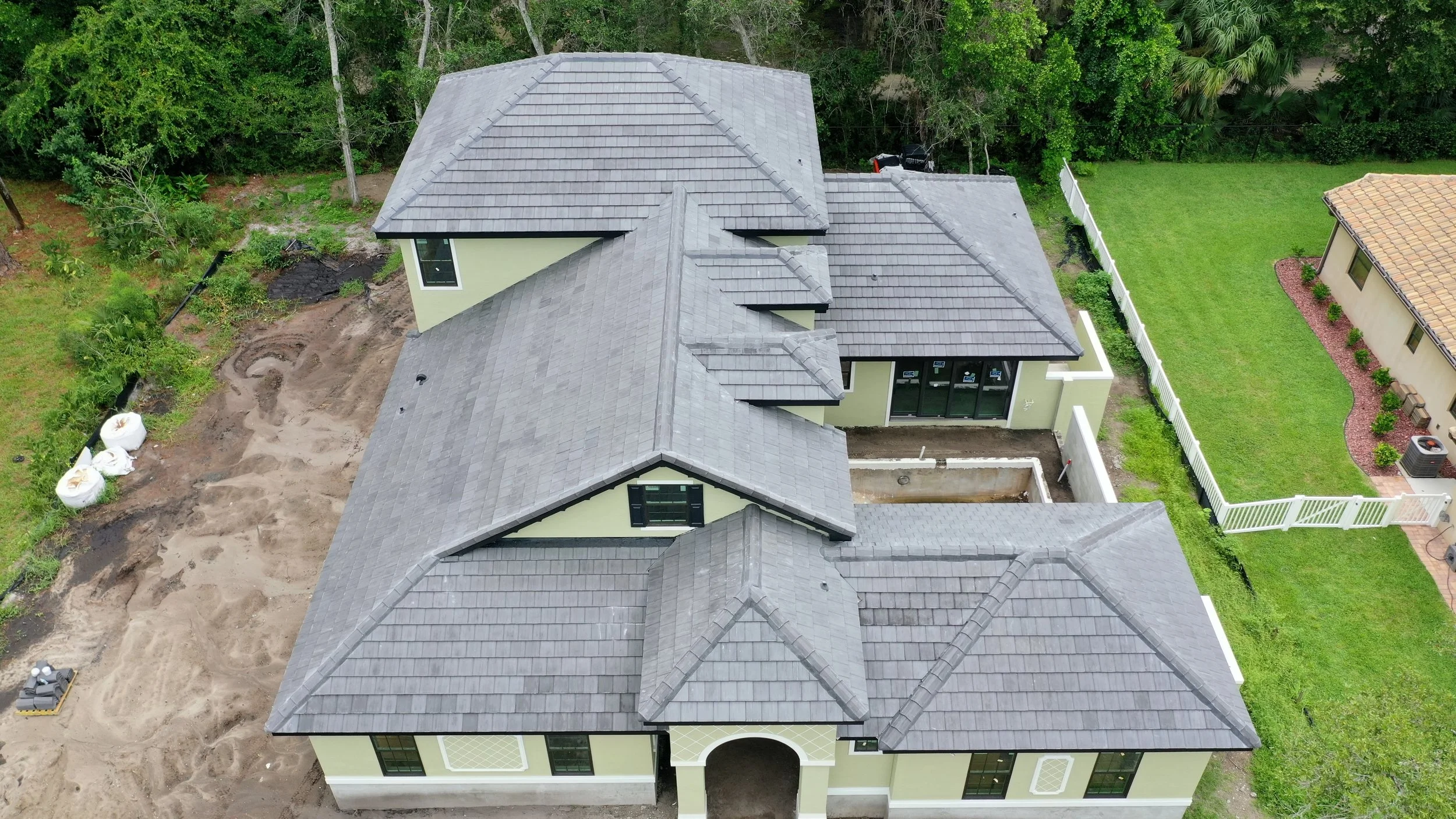 Aerial view of a new house under construction with a light green exterior and gray tiled roof, surrounded by a backyard with a fence, adjacent to a neighboring house and greenery.