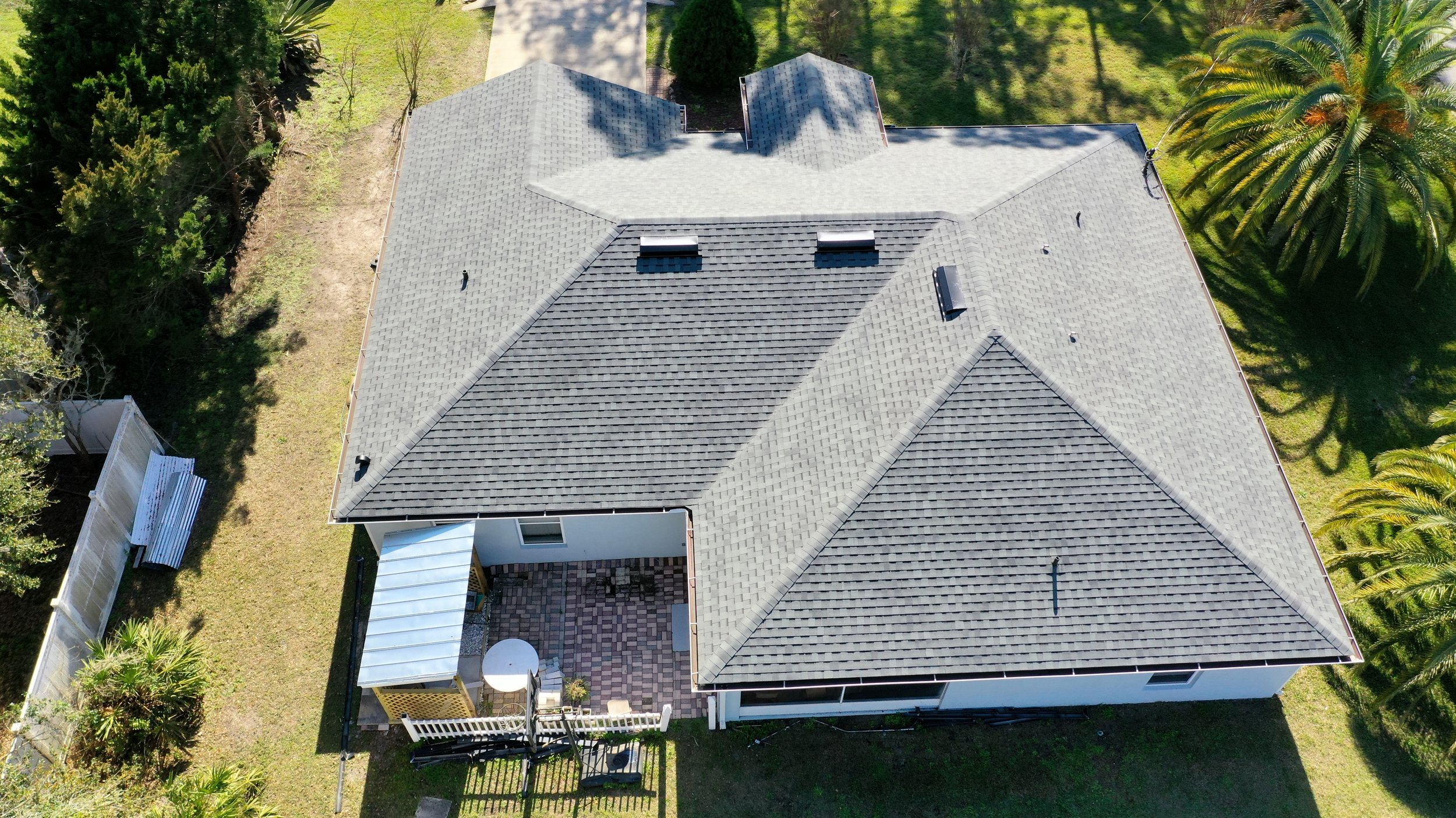 Aerial view of a house with a gray shingle roof surrounded by green trees and lawn, with a fenced backyard patio area.