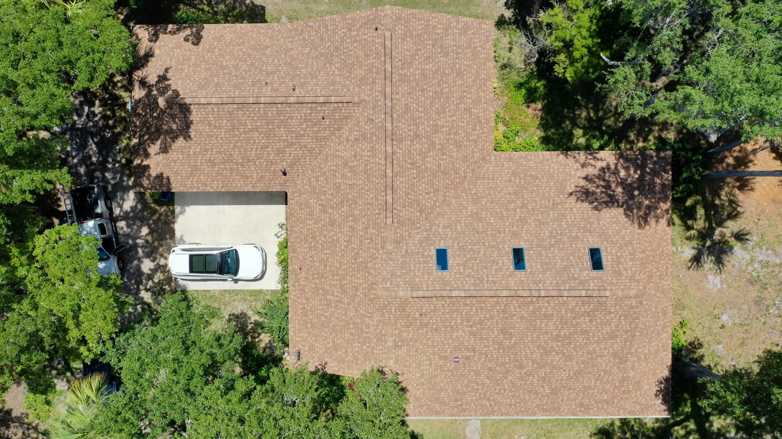 An aerial view of a house with a brown shingled roof, three skylights, and a driveway with one parked cars, surrounded by trees.