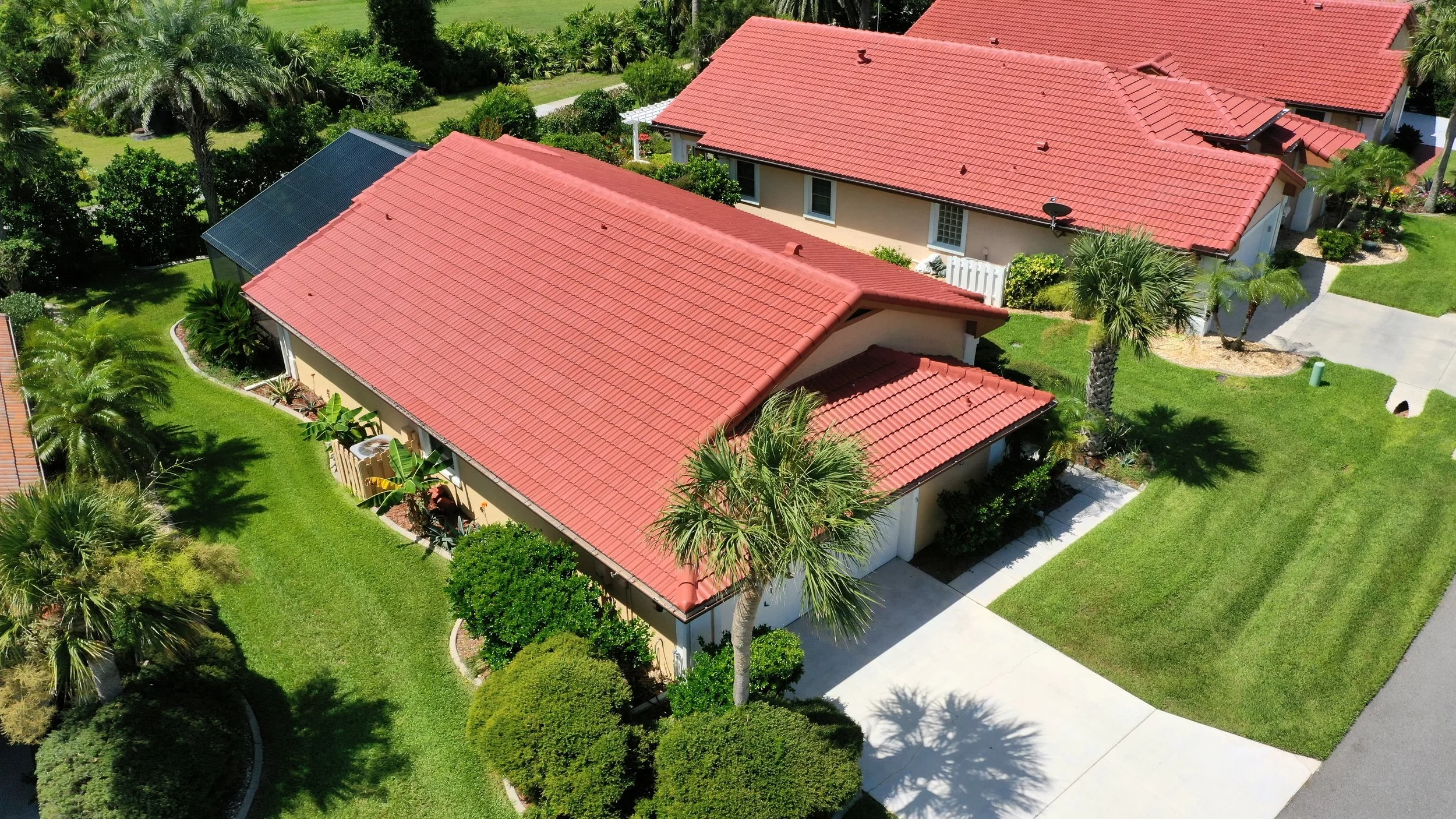 Aerial view of a suburban neighborhood with houses featuring red tile roofs, surrounded by well-maintained green lawns, palm trees, and lush landscaping.