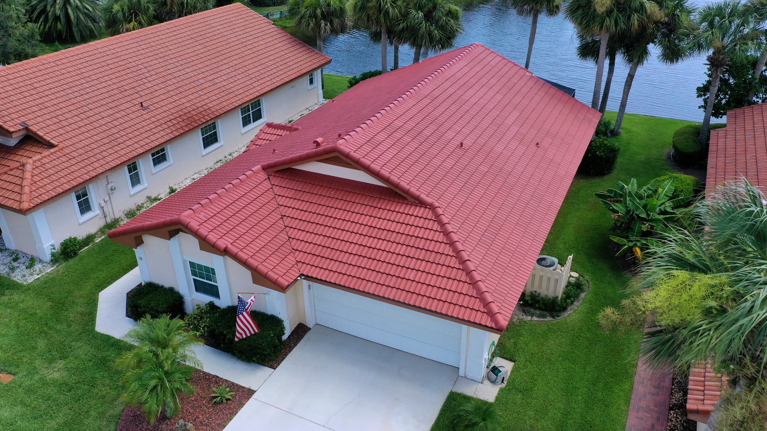 An aerial view of a house with a red tiled roof, white walls, a closed garage door, a small front porch with an American flag, green manicured lawn, palm trees, and a nearby lake or pond.