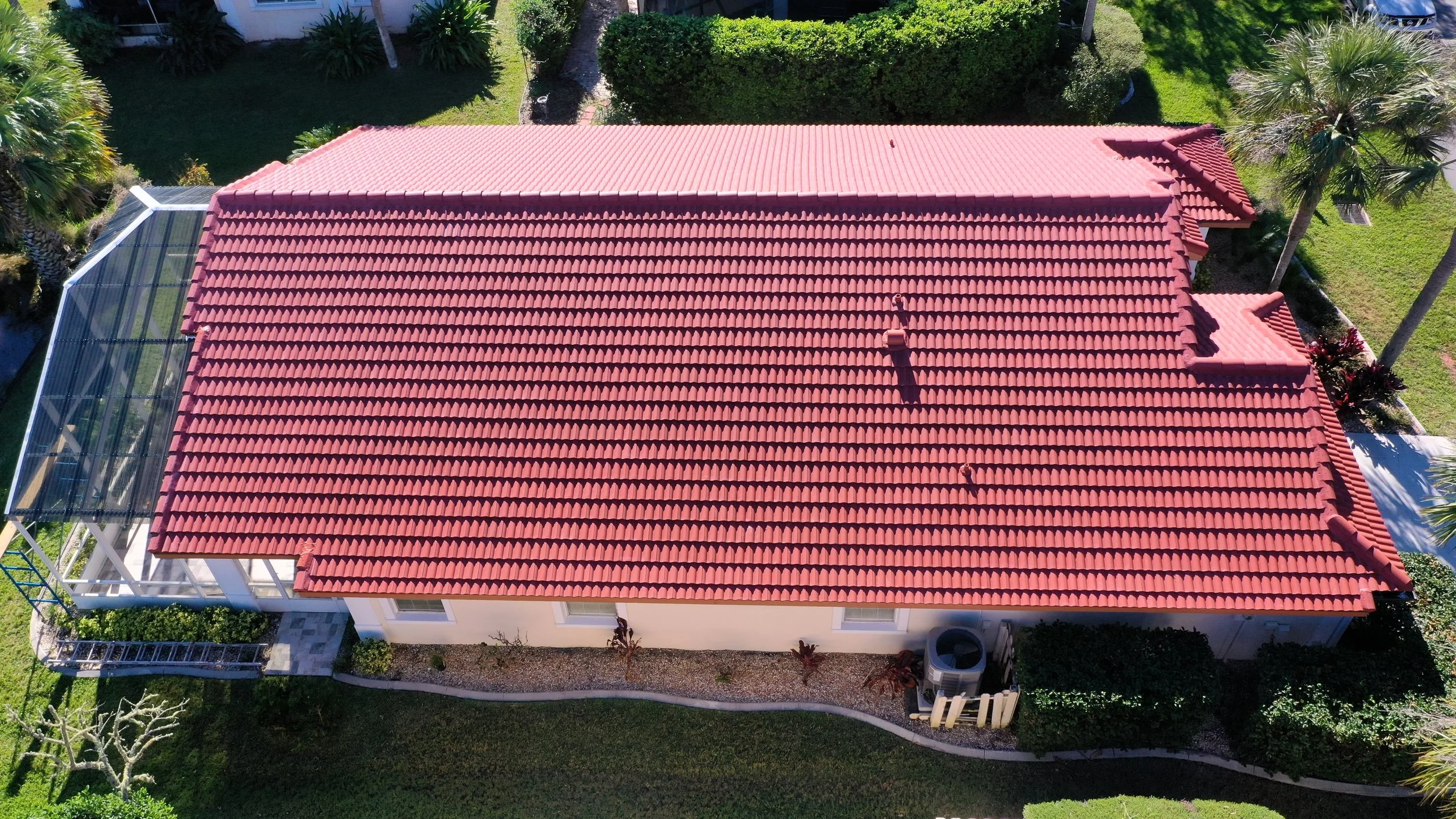 An aerial view of a house with red tiled roof, enclosed porch with glass walls on the left, surrounded by green grass, trees, and bushes.