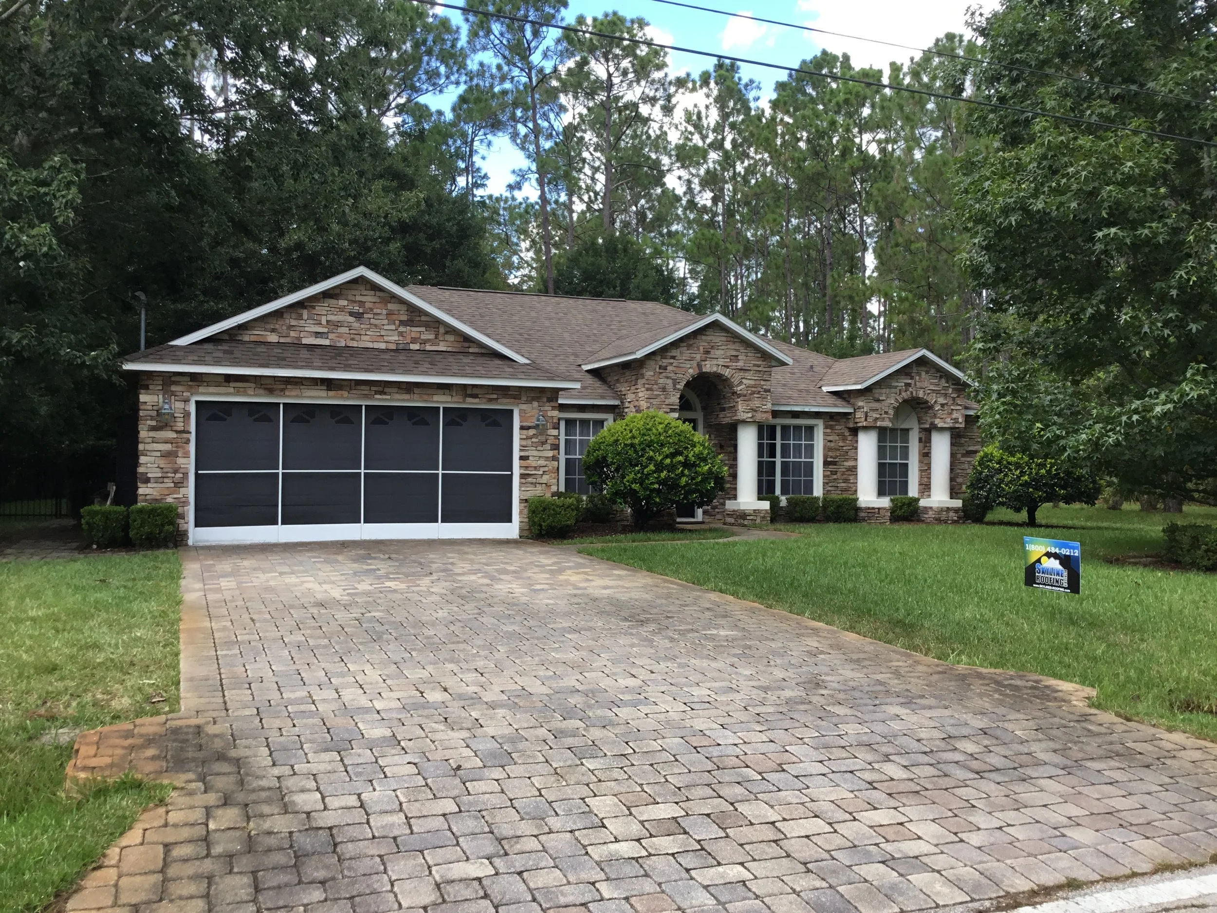 A brick house with a dark garage door, white columns, green bushes, and a lawn, with a paving stone driveway and a house for sale sign in the yard.