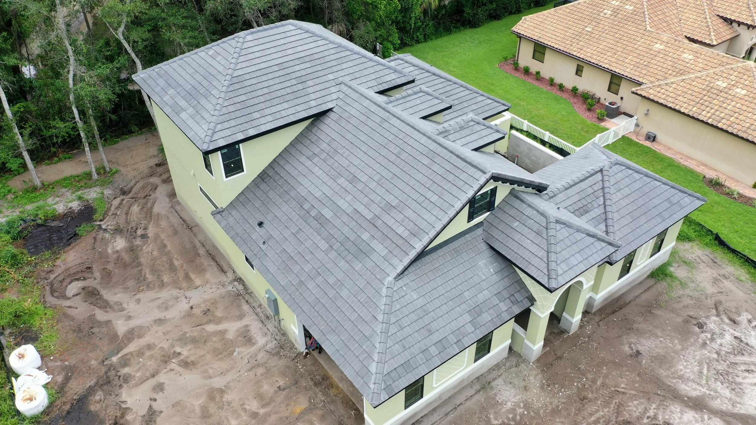 An aerial view of a newly constructed house with gray tiled roof, cream-colored walls, and multiple roof sections. The area around the house is under construction with dirt and no landscaping yet. To the right are neighboring houses with tile roofs a