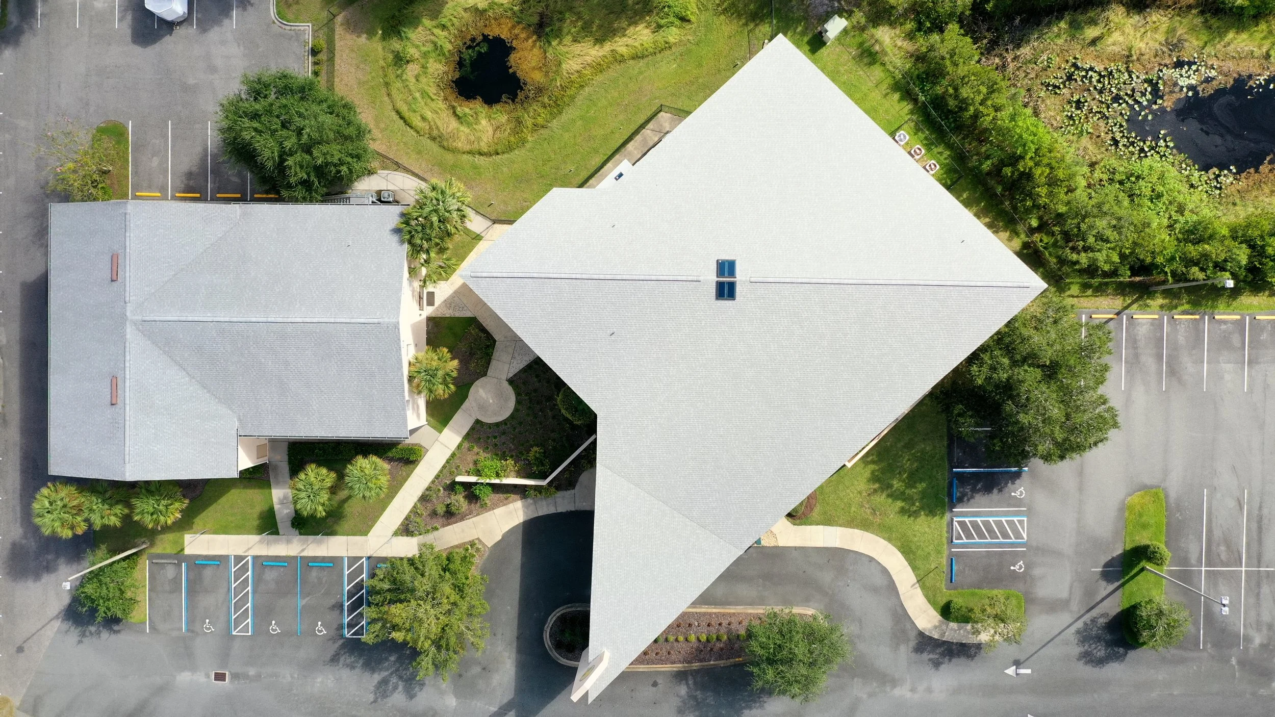 Aerial view of a building with a light-colored roof, parking lots, greenery, and surrounding landscape.