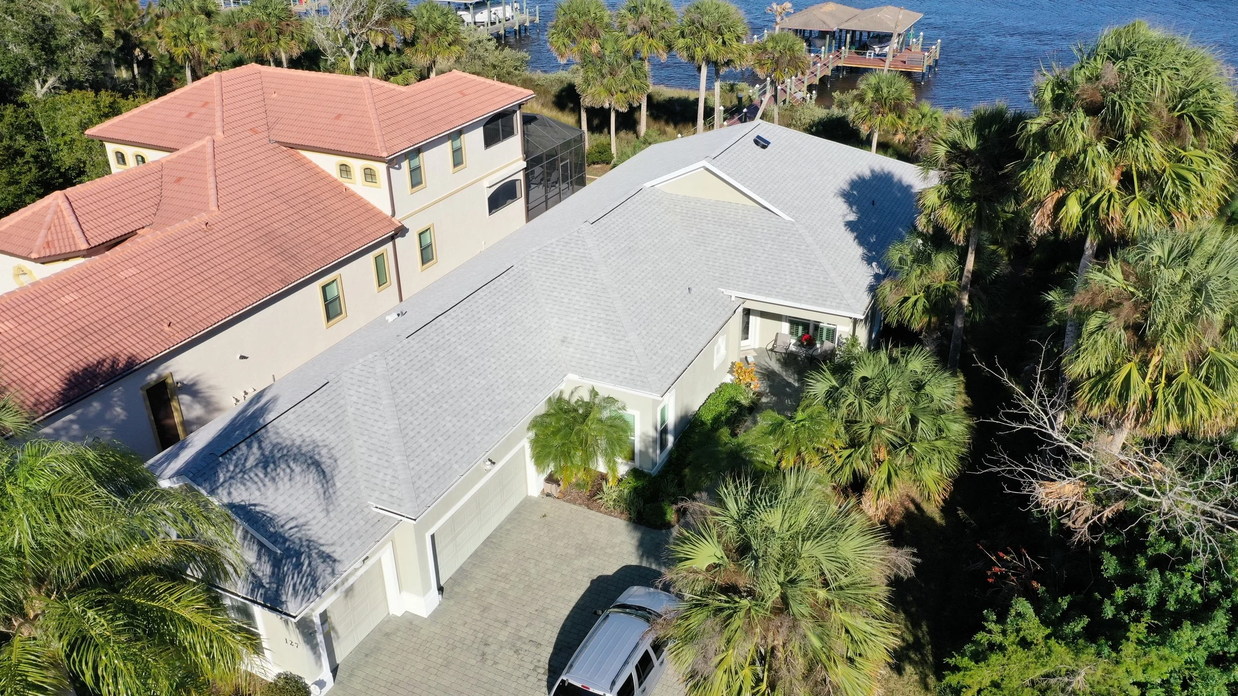 Aerial view of modern house with a gray roof, driveway with a silver car, surrounded by lush green palm trees, with a waterfront and dock in the background.