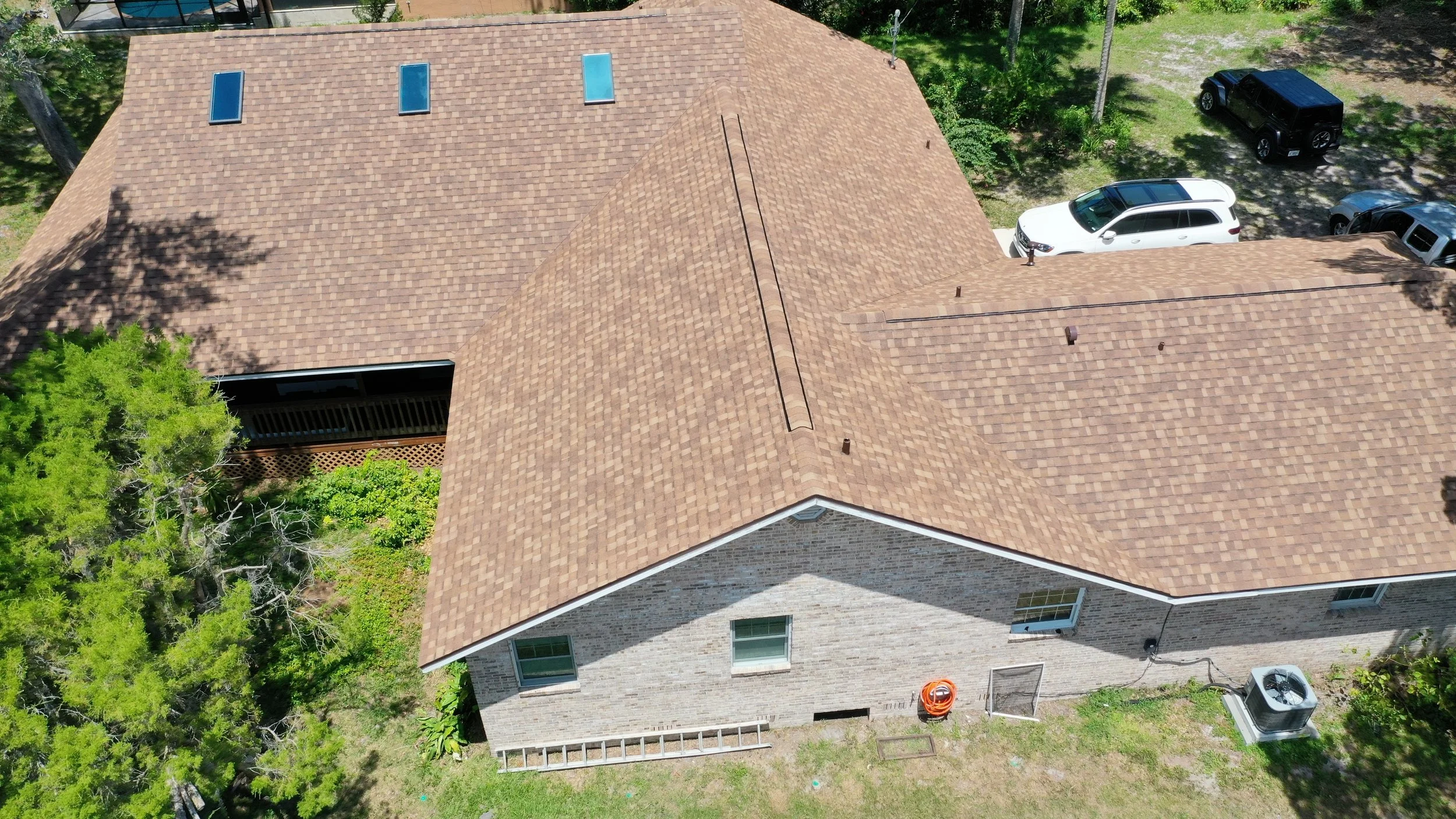 Aerial view of a house with a brown shingle roof, multiple windows, and a backyard with some green trees, a ladder, an orange hose, and an air conditioning unit. A driveway with three parked vehicles is visible to the right.
