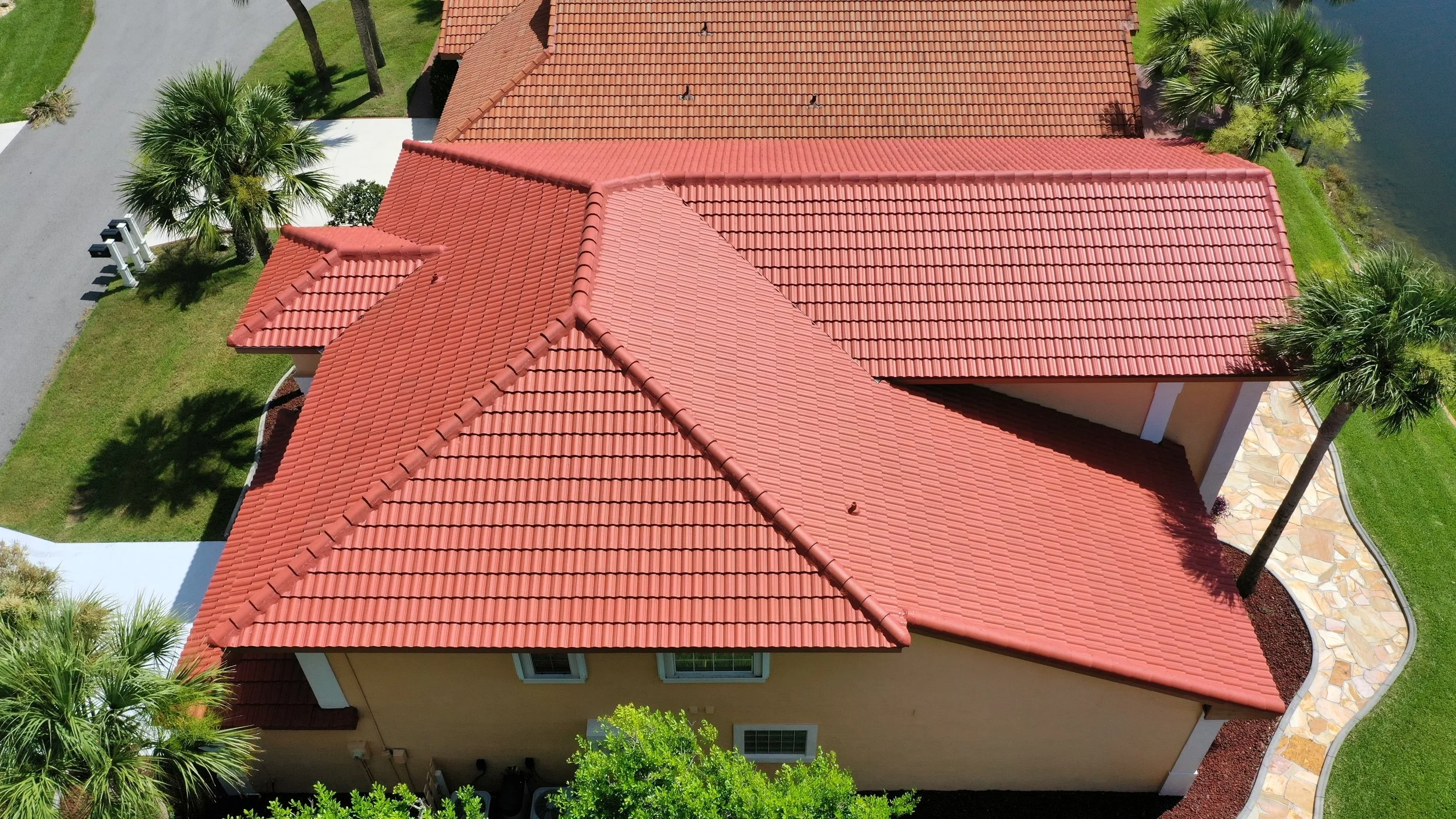 Aerial view of a house with red tiled roof, palm trees, a walkway, green lawn, and adjacent water body.