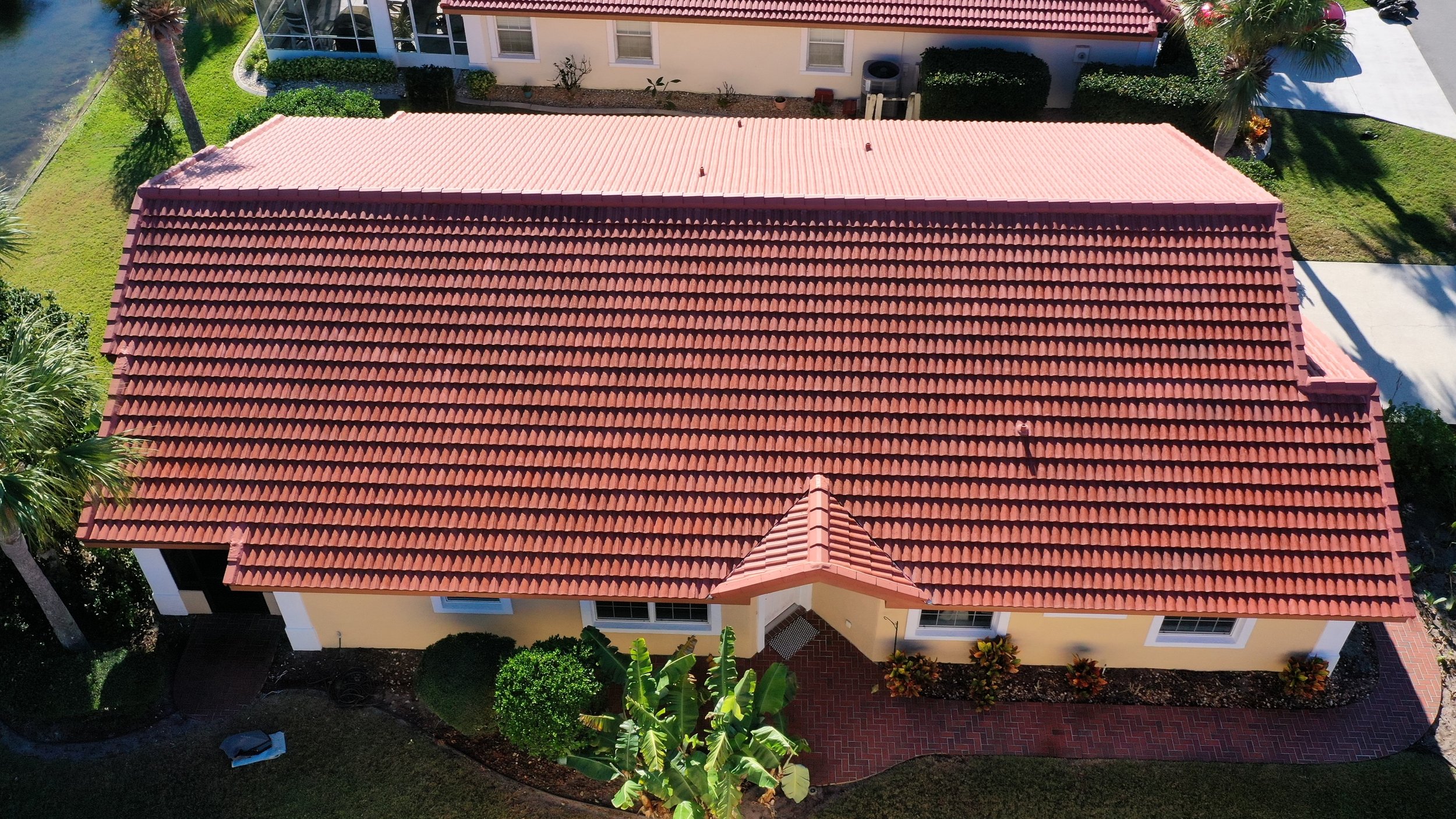 Aerial view of a house with a red tiled roof, yellow exterior walls, surrounded by green trees, shrubs, and a lawn, with a pathway leading to the entrance.