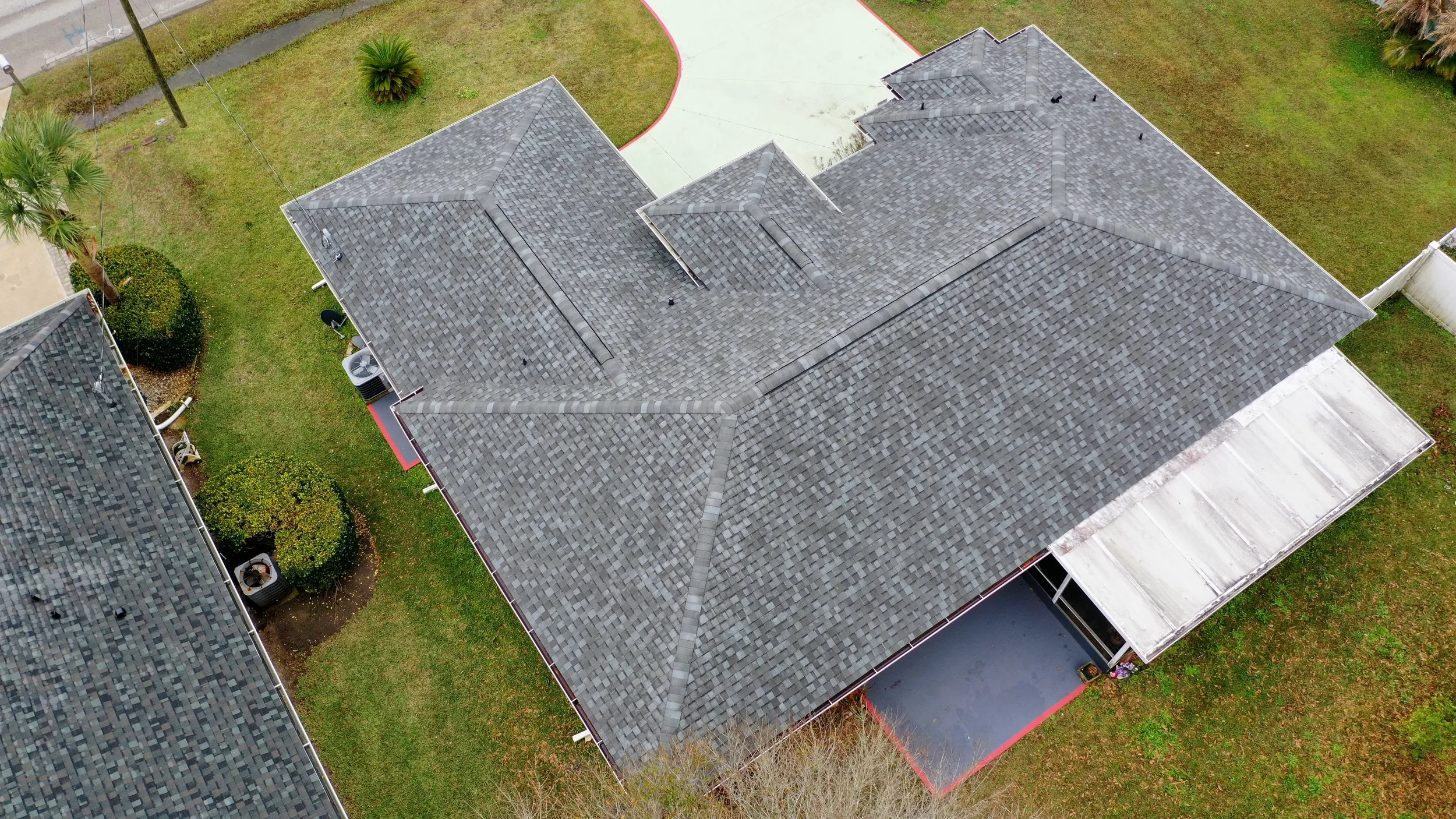 Aerial view of a house with a gray shingle roof, surrounded by a grassy yard with trees and bushes, and neighboring houses.