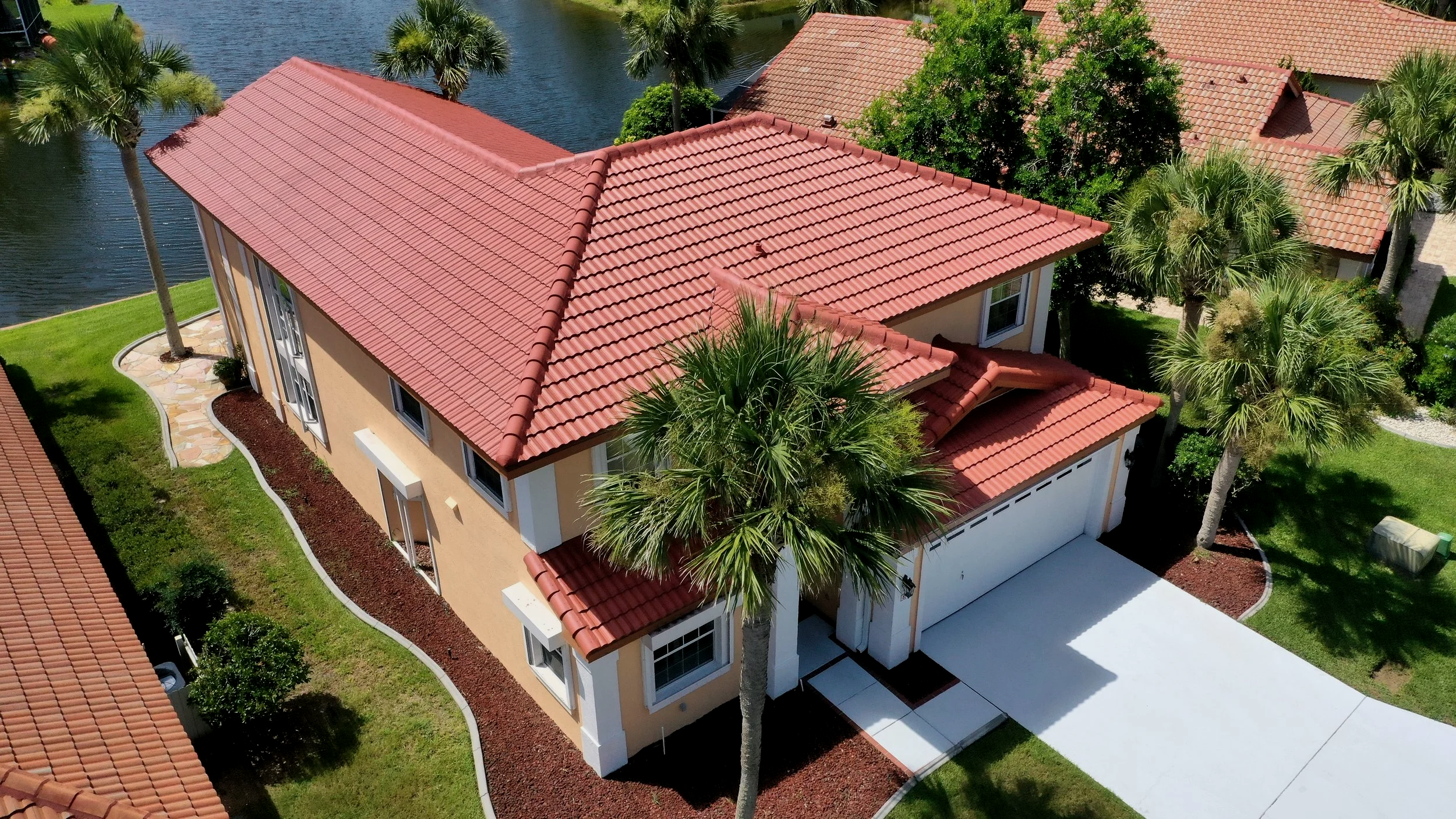 Aerial view of a modern house with a red tile roof, surrounded by palm trees, green lawn, and a nearby water body.