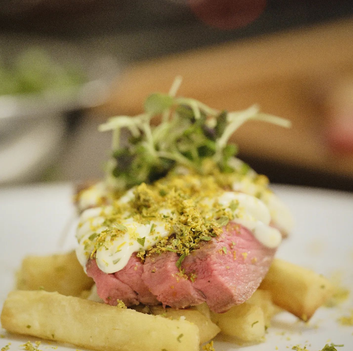 Gourmet dish with a pink meat patty, topped with sauce and herbs, served over fries.
